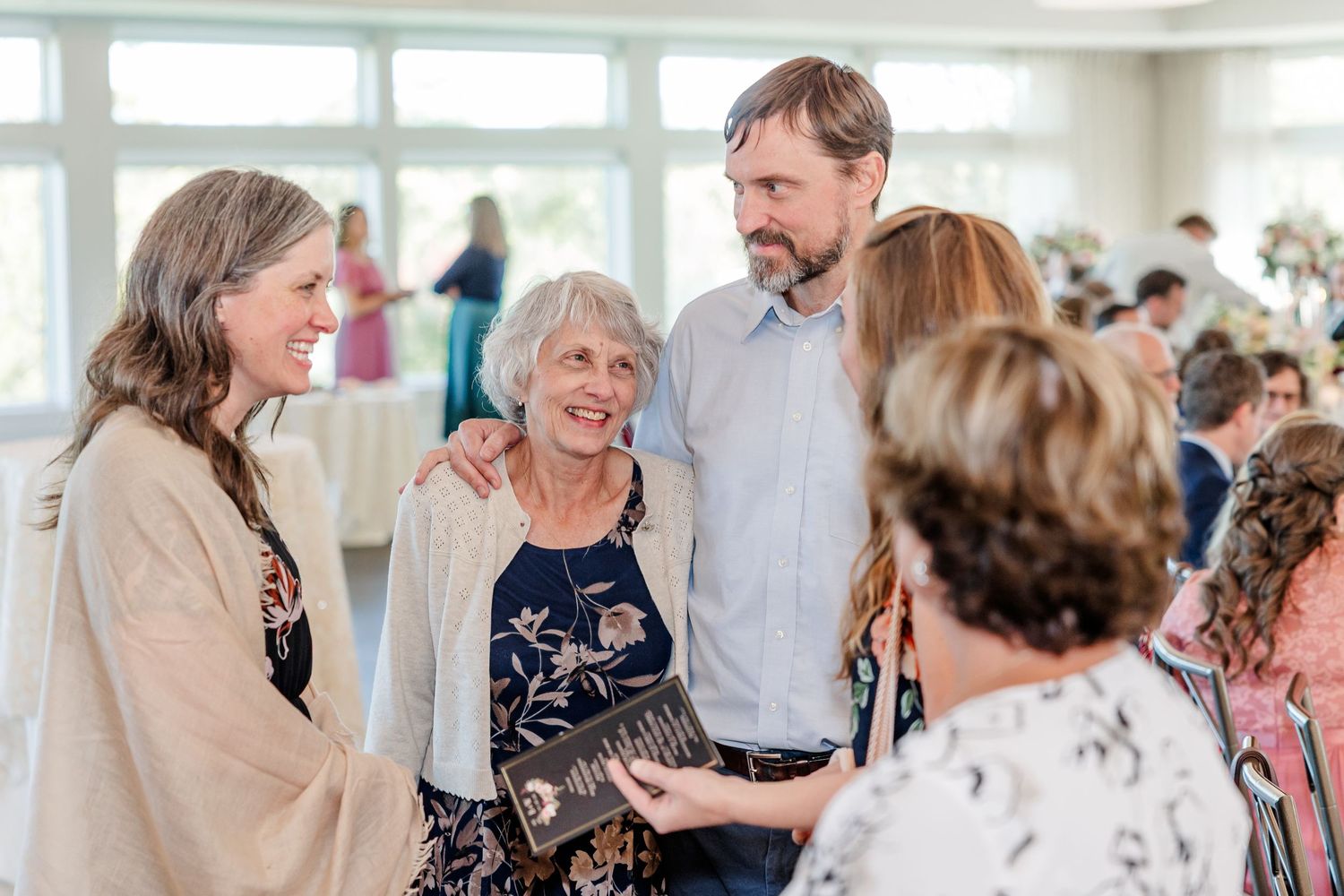 Smiling older woman holding a framed plaque is embraced by a tall bearded man and a woman at a lively indoor reception with guests.