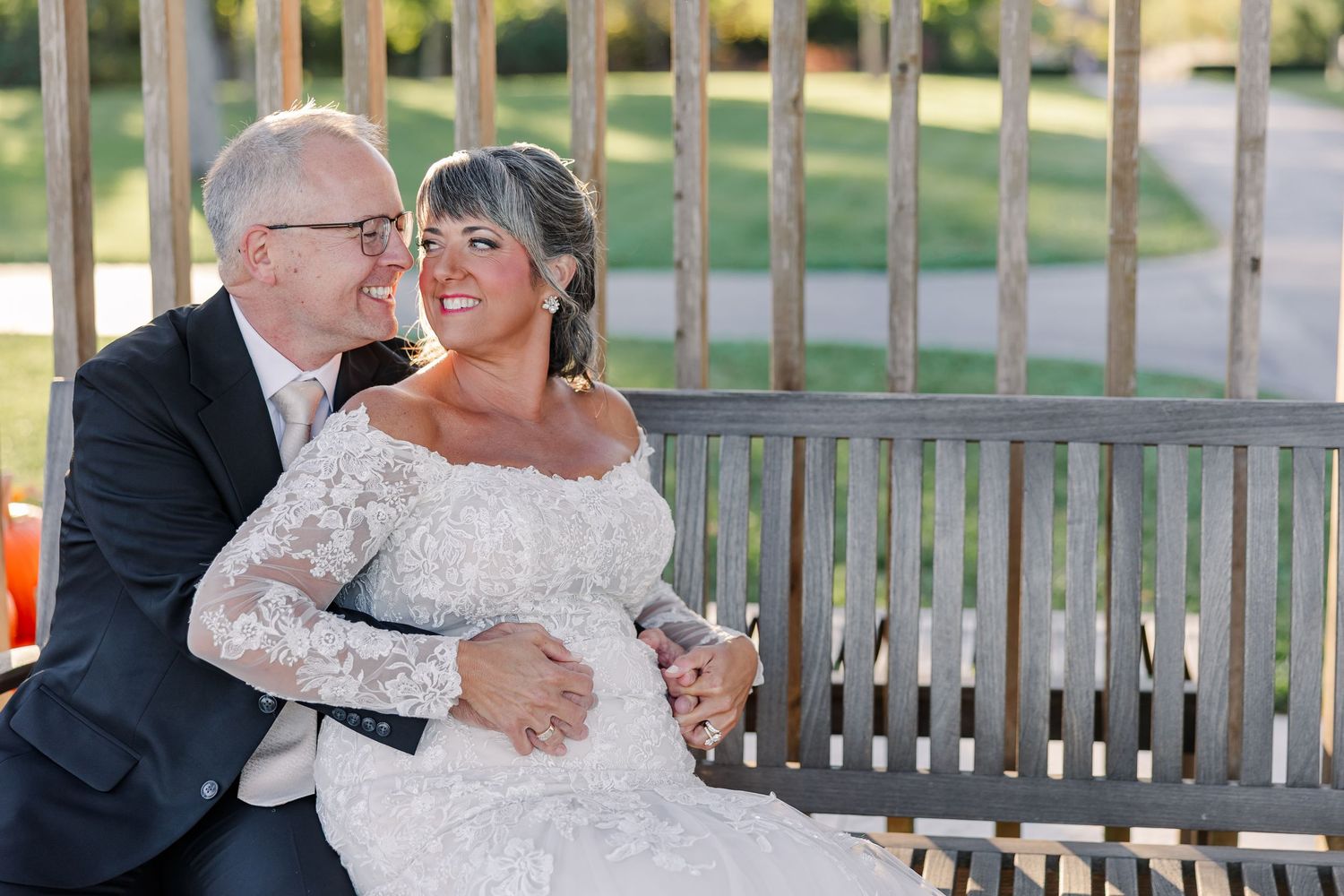 Smiling bride in a lace wedding gown sits close to her groom in a dark suit on a wooden garden bench, his hand resting on her waist.