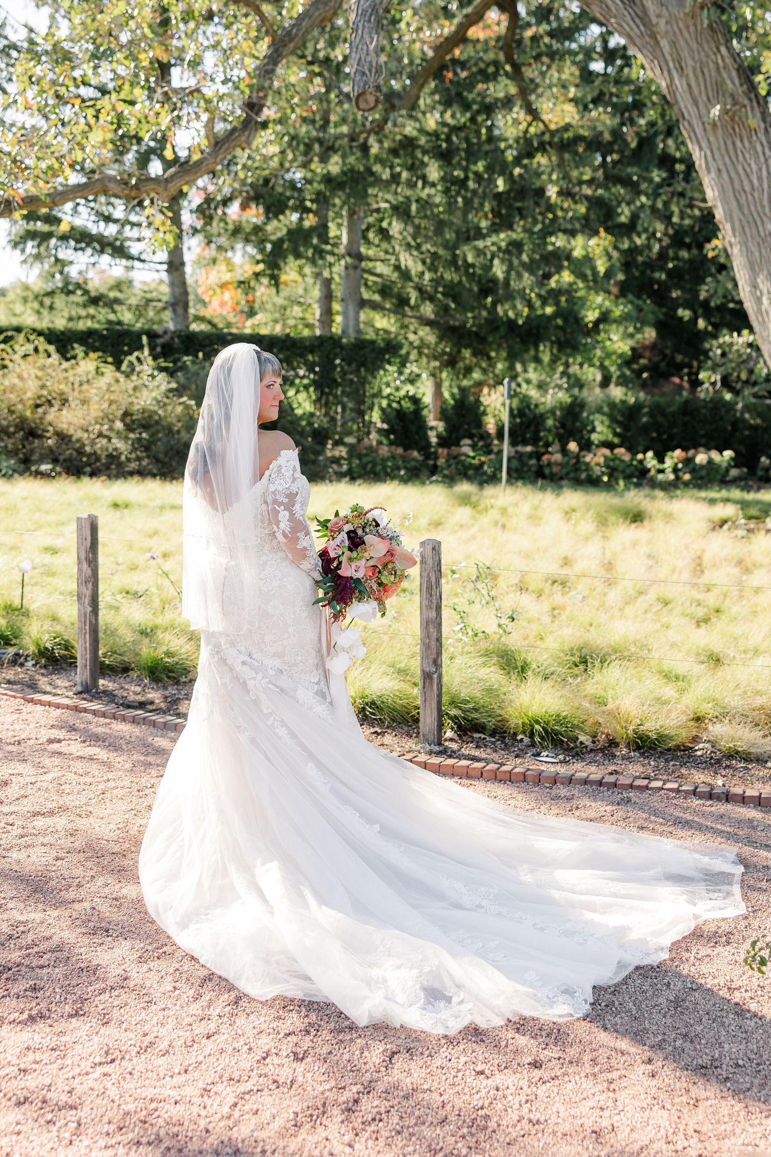 Bride in a lace wedding gown with a long veil and train holds a colorful bouquet while standing in a sunny garden.