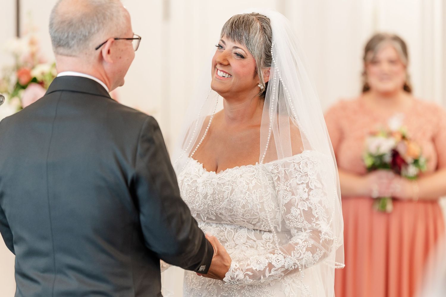 Smiling bride in a lace gown and veil holds hands with the groom at the altar while a bridesmaid in coral holds a bouquet.