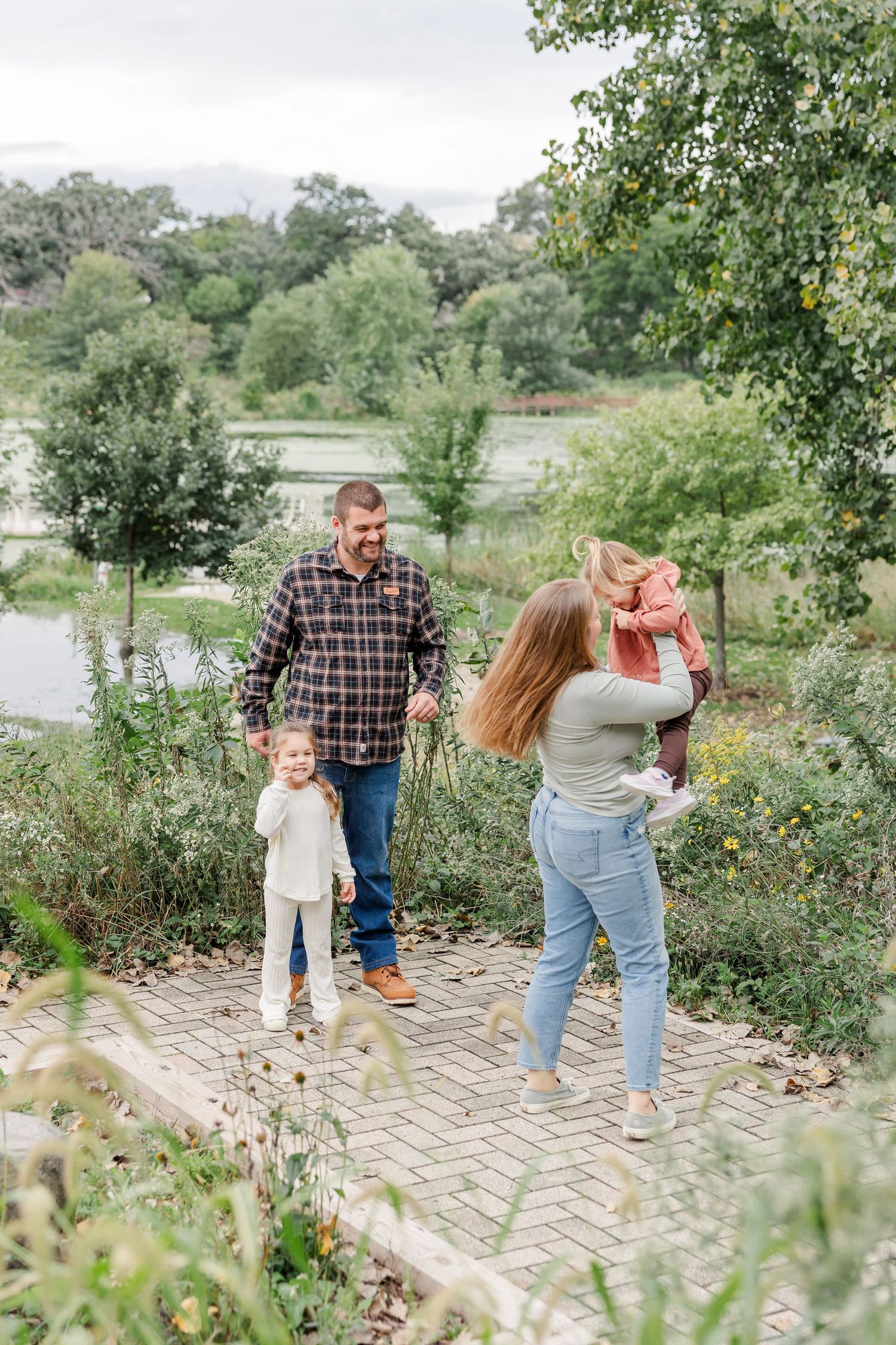 A happy family of four enjoys a sunny day on a brick path surrounded by lush greenery near a pond.