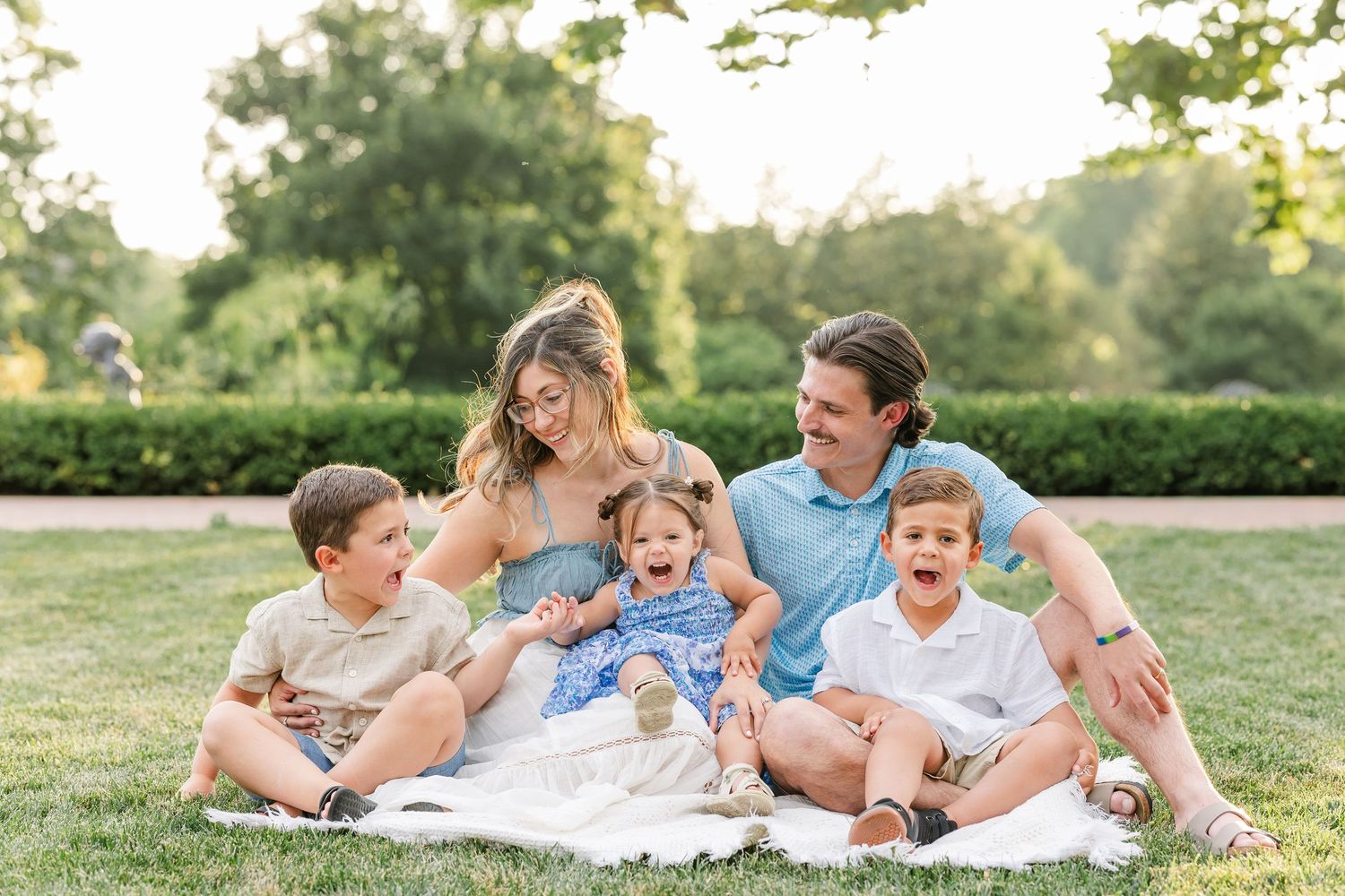 Happy family of five sitting on grass in a park, laughing together on a sunny day.