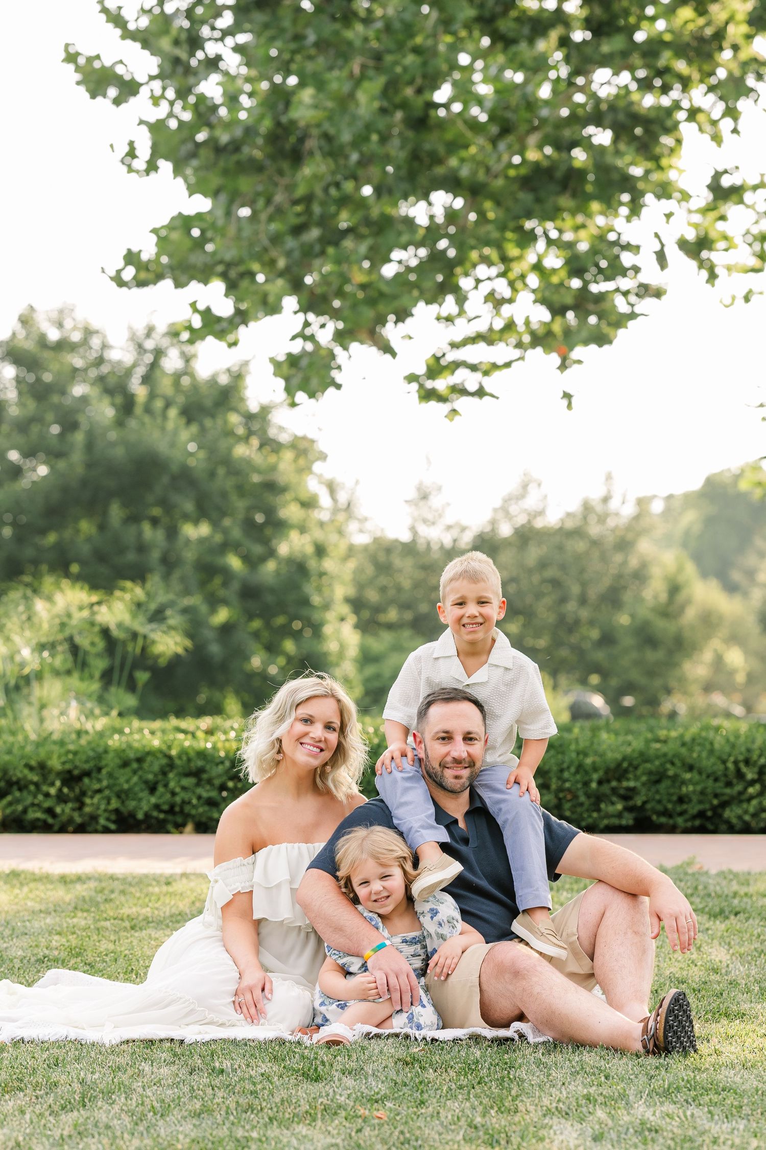 A happy family of four poses on green grass outdoors, with lush trees in the background on a sunny day.