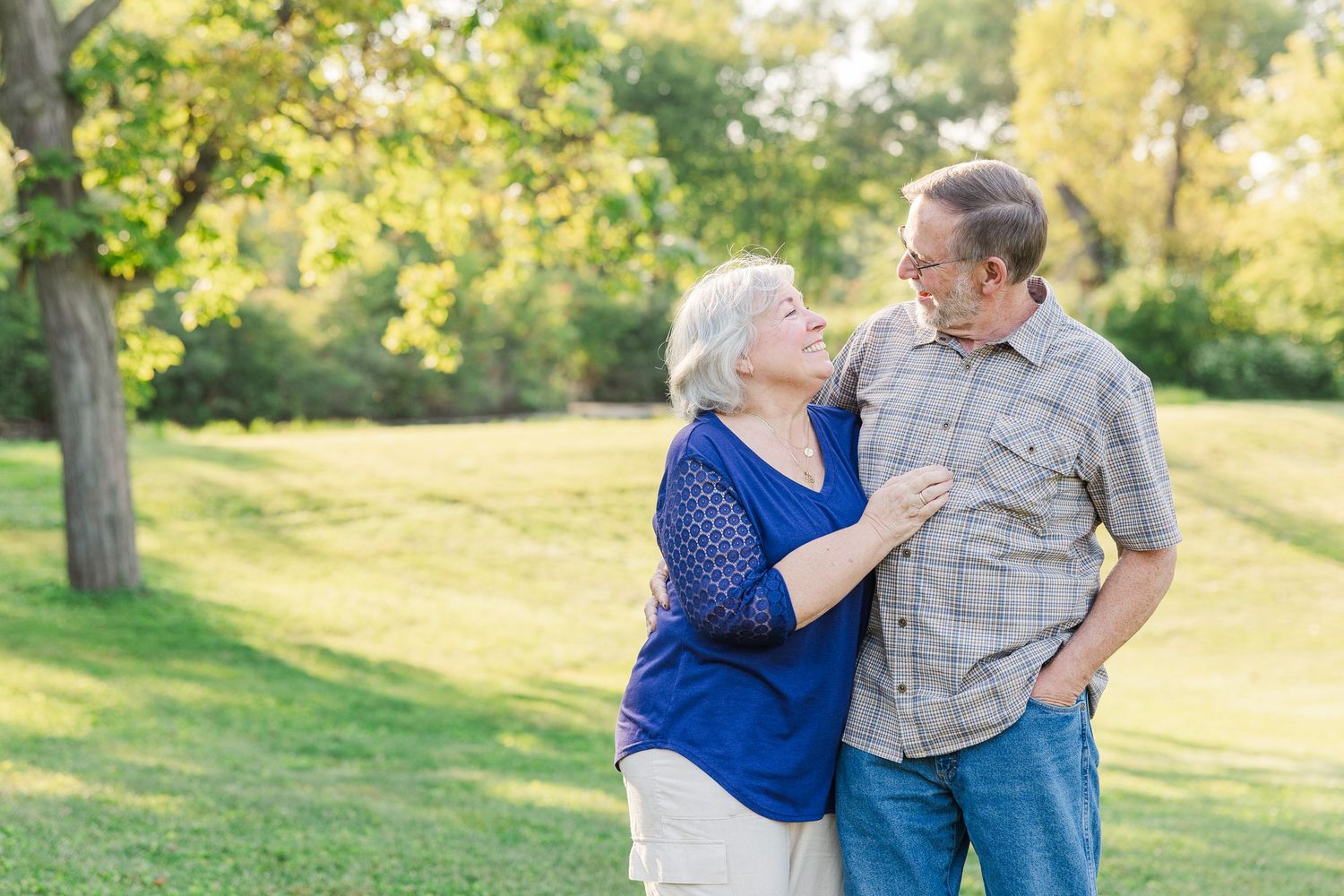 Couple embraces in a sunny park setting during golden hour amidst trees and lush grass.Sorg family portraits at Shamrock Hills Park in Cryst