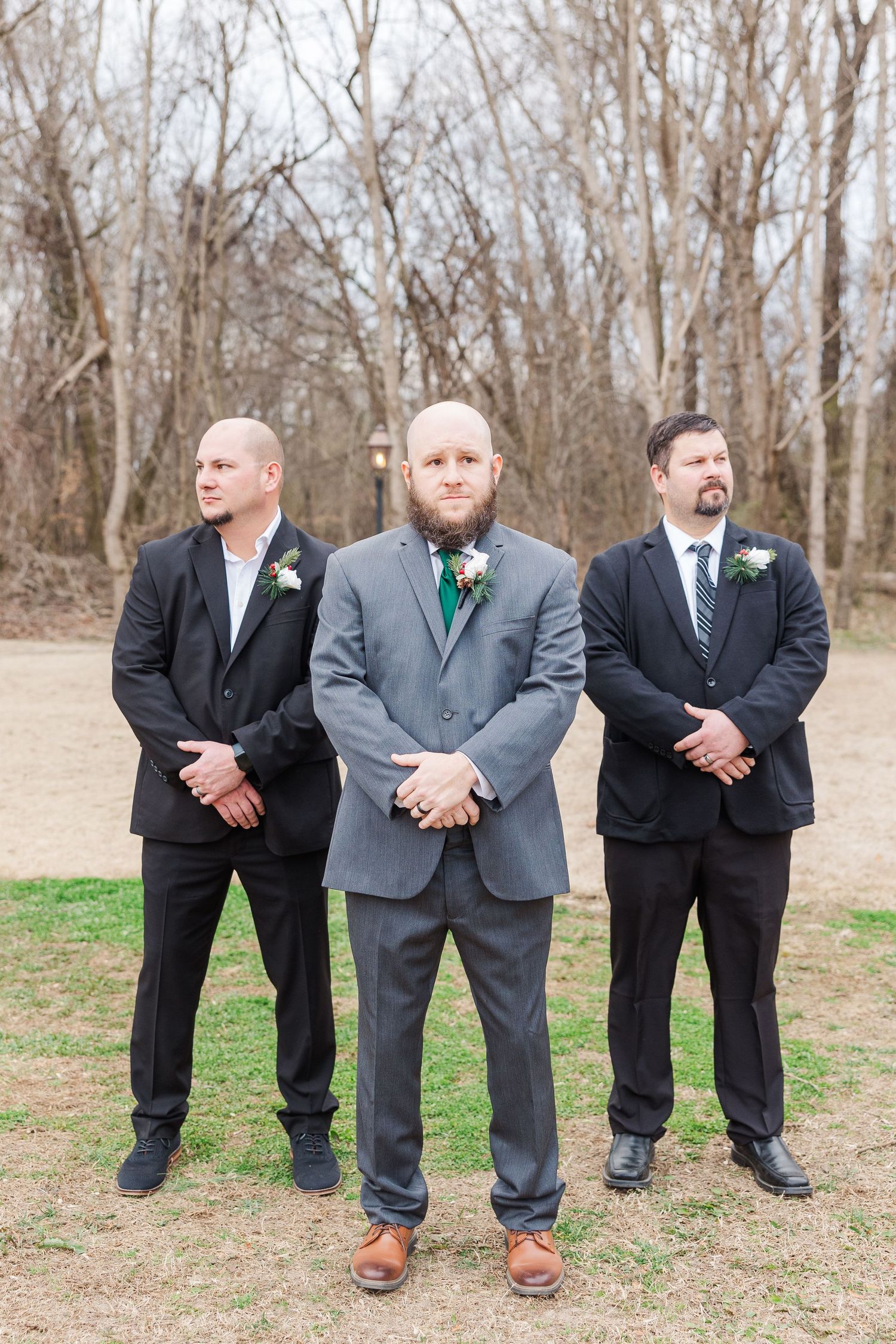 Three groomsmen in formal suits pose confidently outdoors at a wedding.
