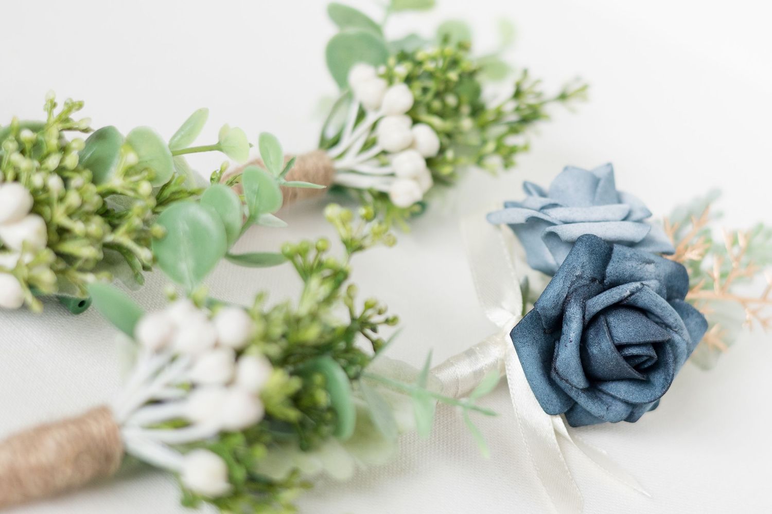 Wedding boutonniere and corsage with blue roses and greenery wrapped in twine on white background.