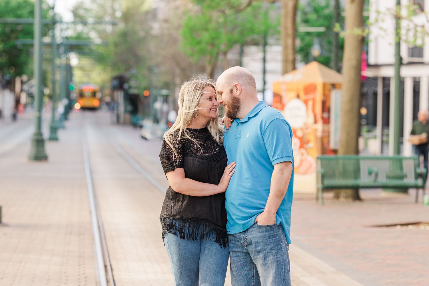 A couple shares an intimate moment on a city street with green trees and lampposts.