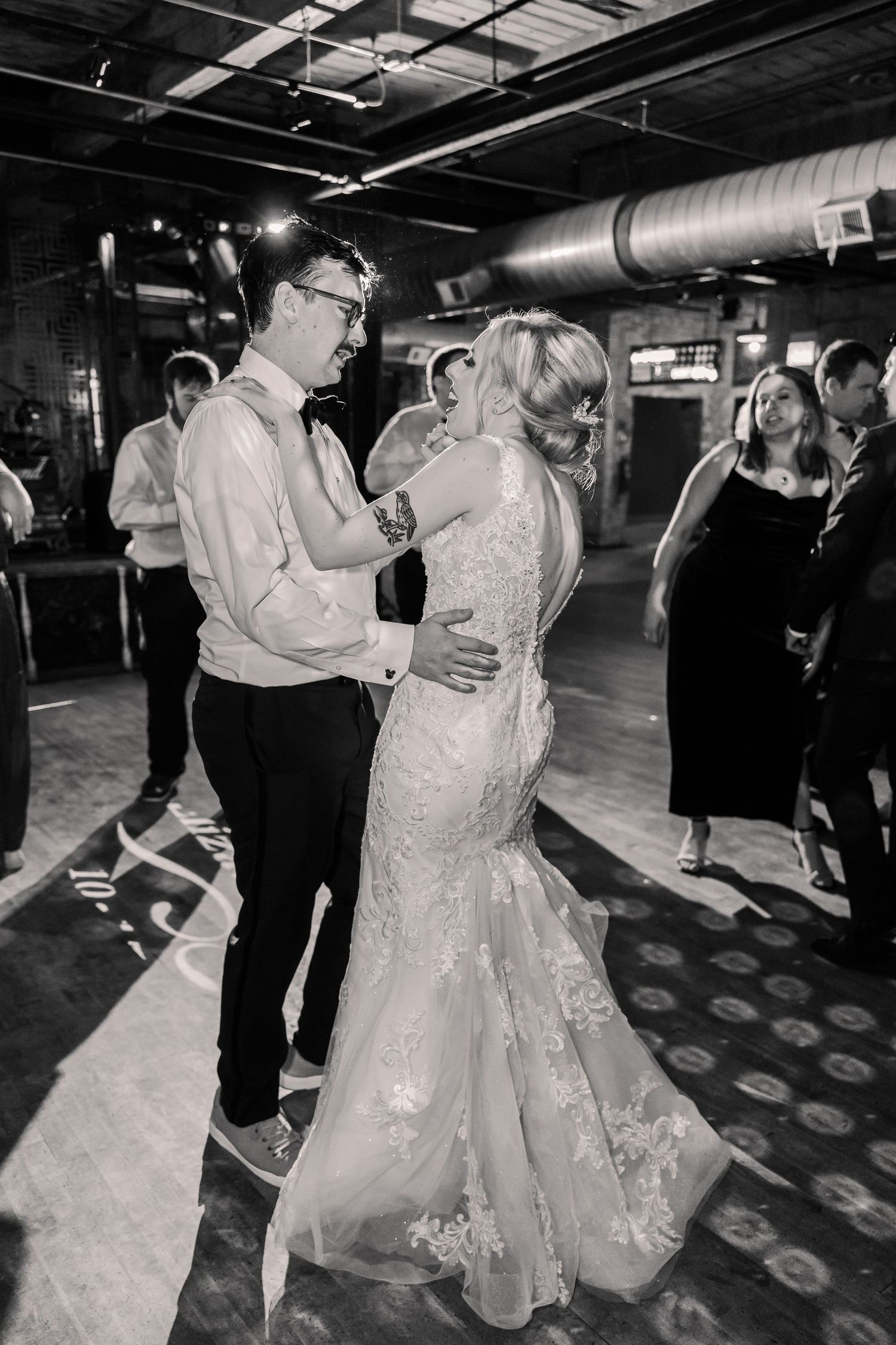 A black and white photo of a couple dancing together in a reception venue with industrial-style ductwork.