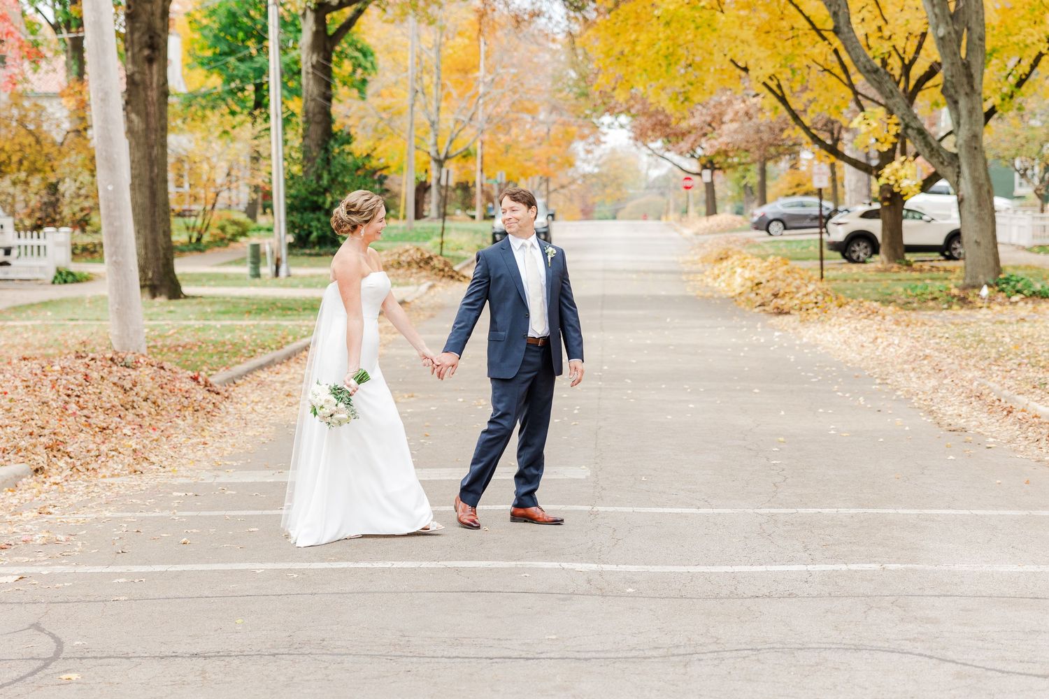 Wedding couple strolling on leaf-covered street during fall photoshoot.