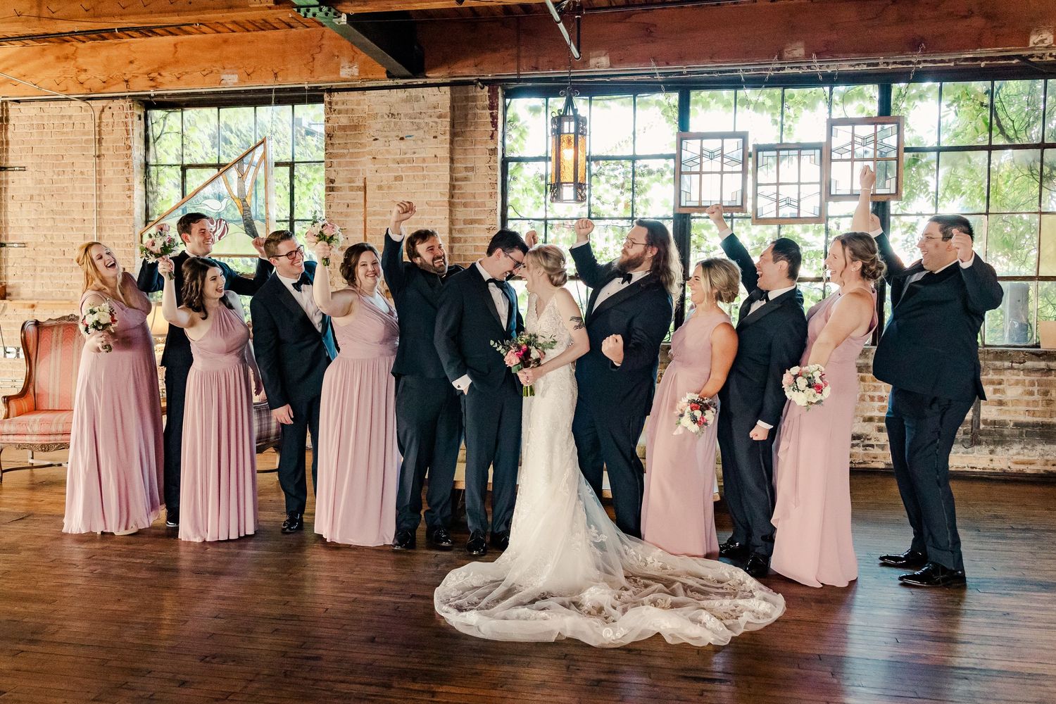 Wedding party celebrates with animated poses in rustic brick venue with natural lighting.