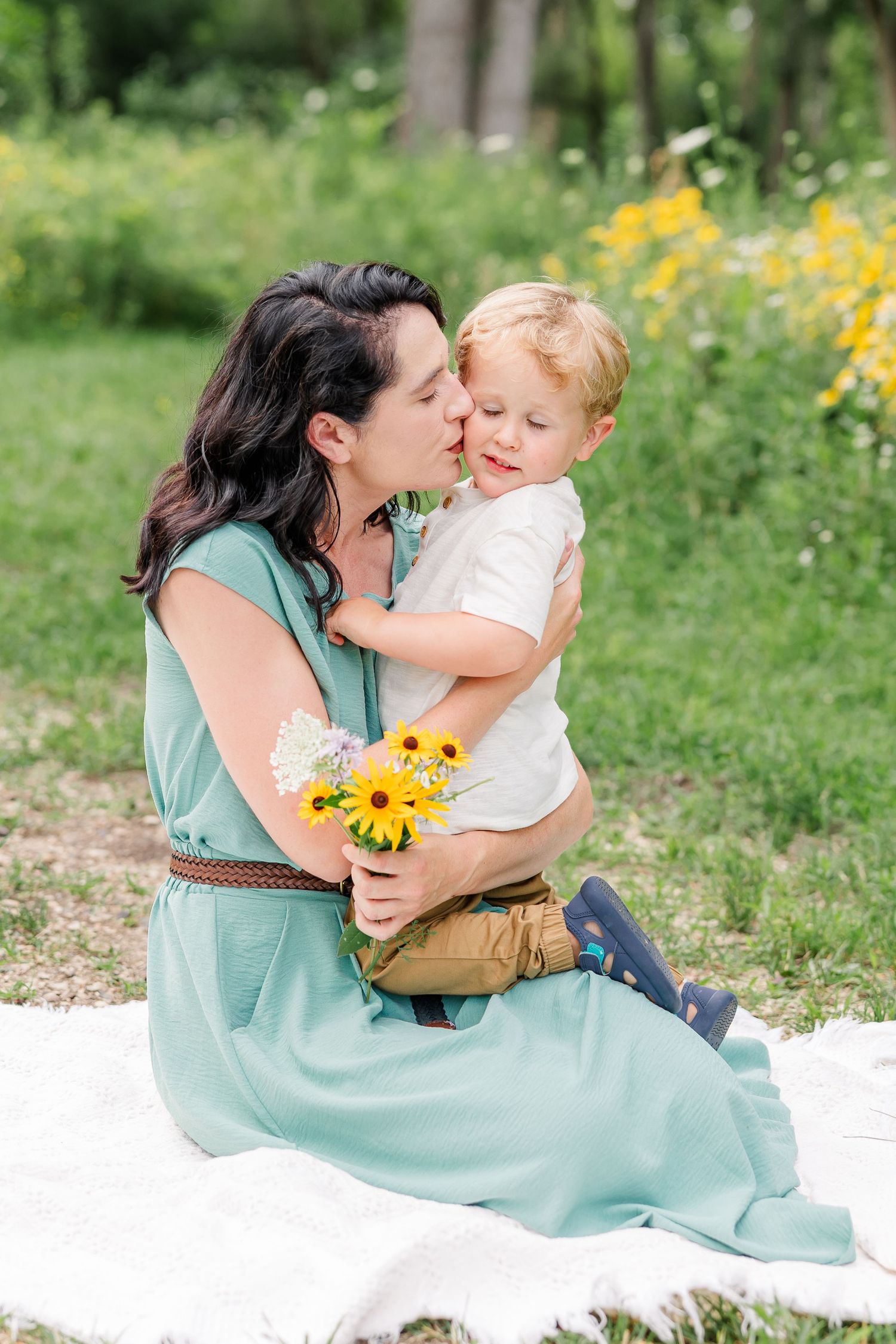 A mother in a teal dress kisses her toddler son while holding yellow wildflowers on a blanket in a green park.