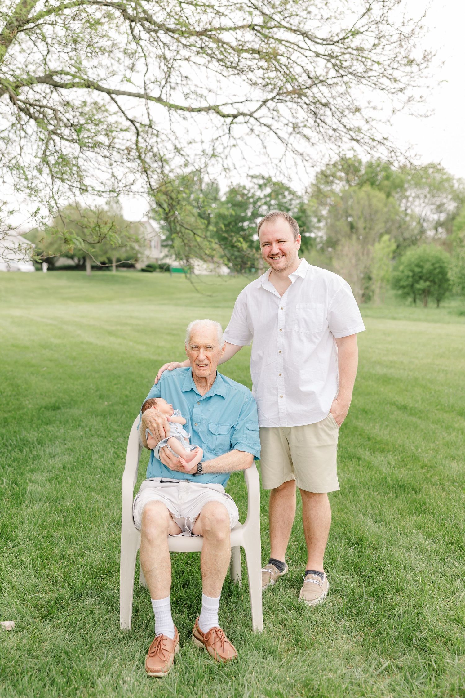 Elderly grandfather seated holding newborn baby with adult son standing beside him outdoors.