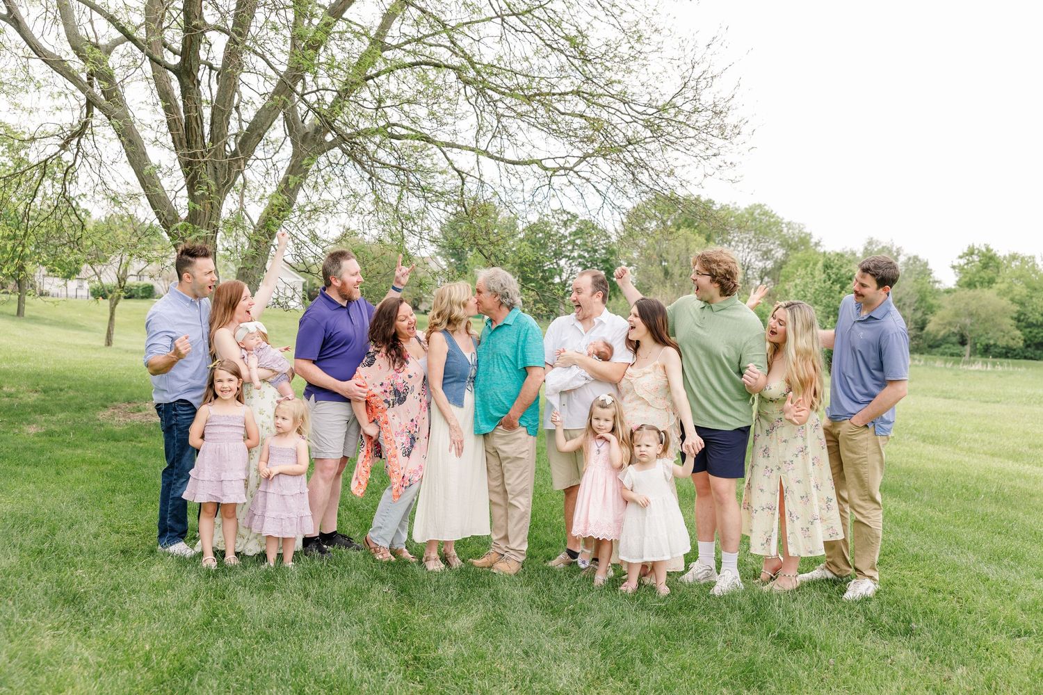 Large multi-generational family gathering outdoors on green lawn beneath a blooming spring tree.