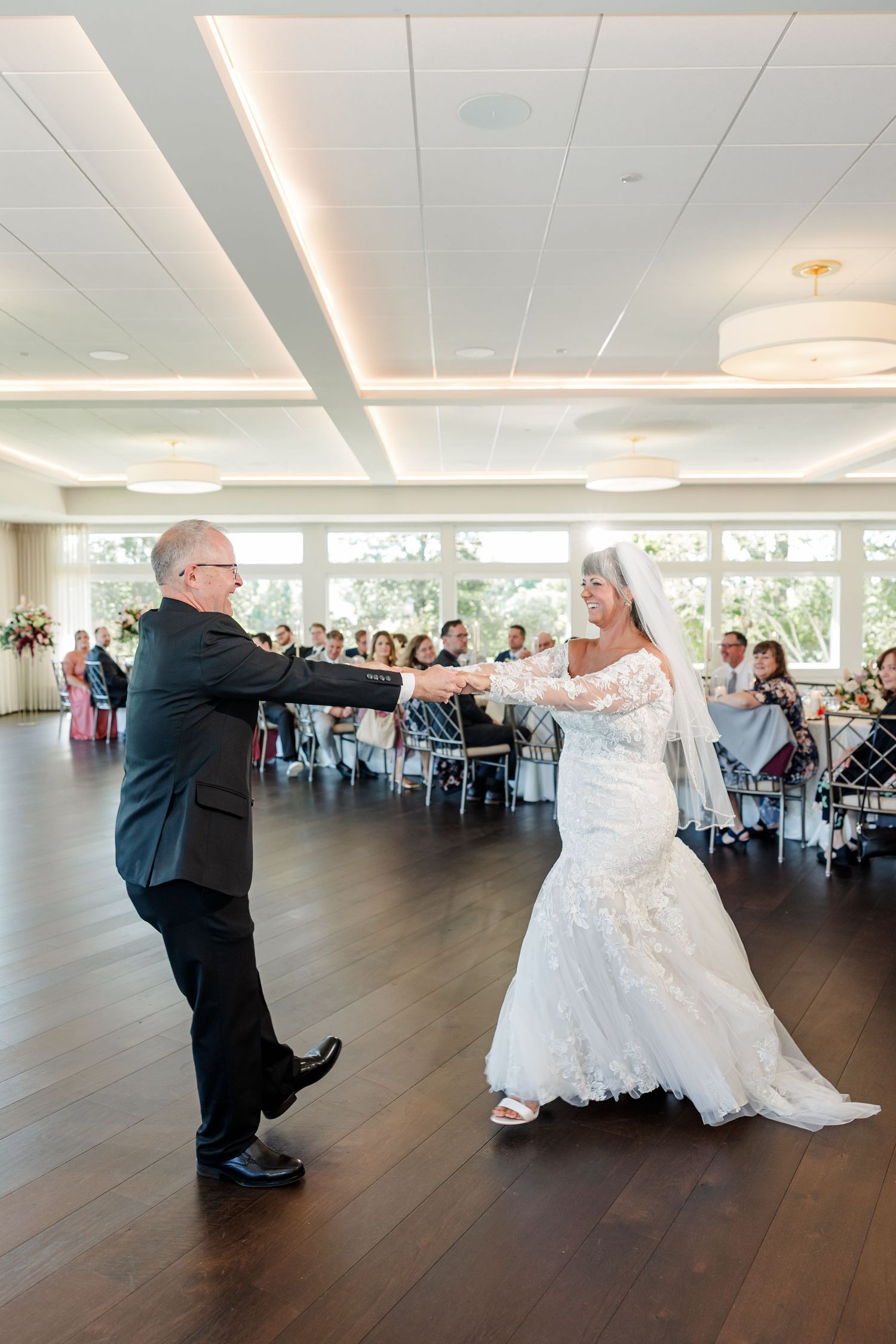 A bride in a lace mermaid gown and veil dances with a man in a dark suit on a wood floor as wedding guests watch from tables.