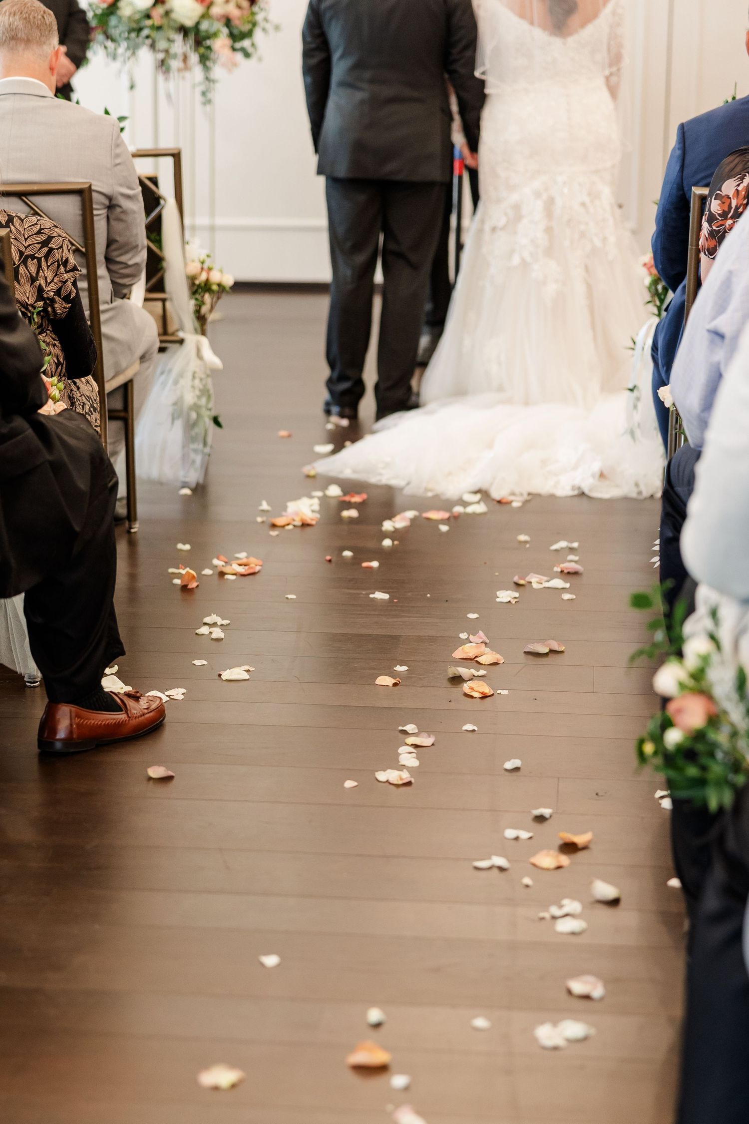 Rose petals scattered along a wooden aisle floor as a bride in a flowing white gown stands at the front of a wedding ceremony.