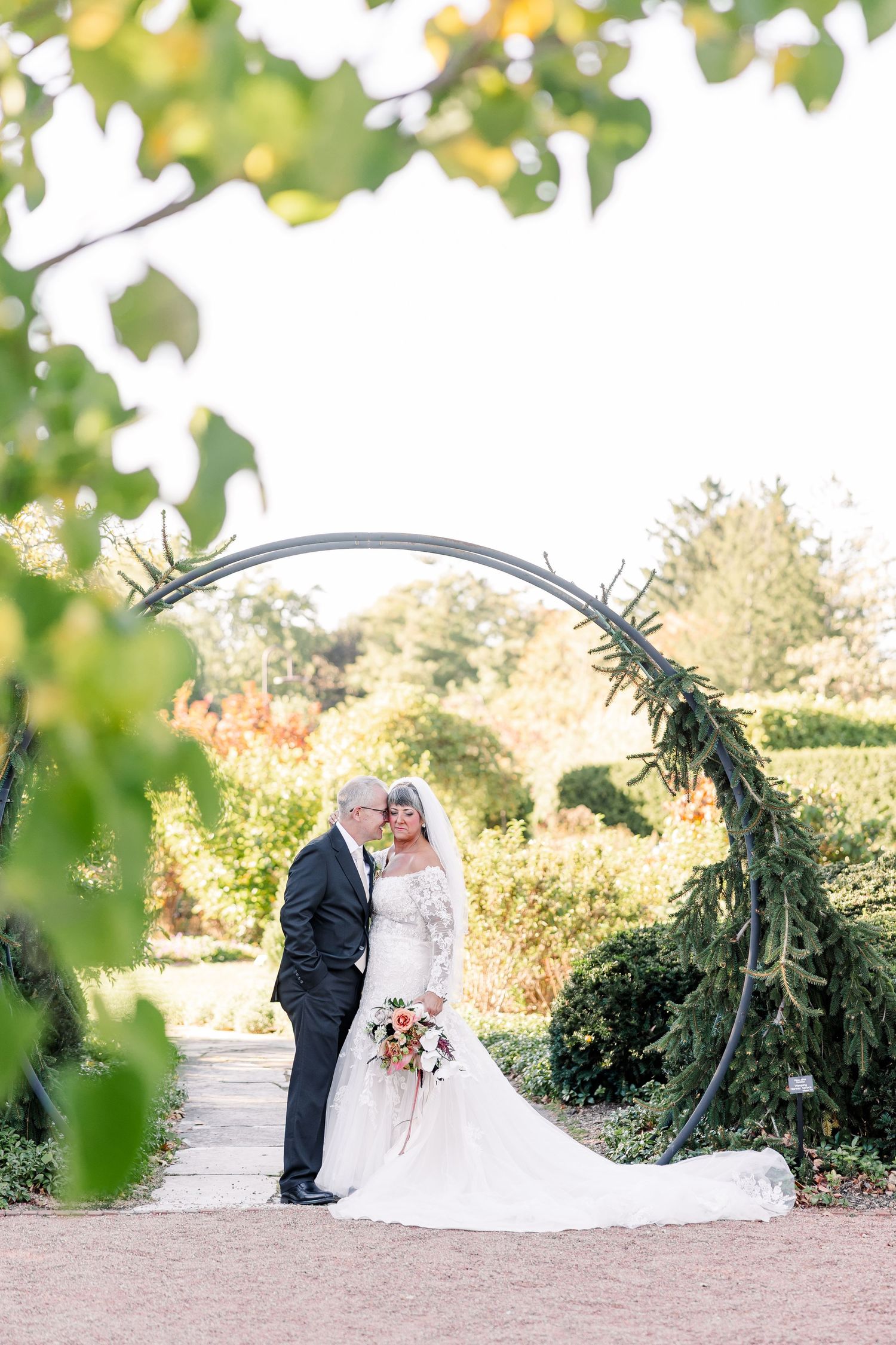 Bride in a lace gown and groom in a dark suit embrace beneath a circular evergreen wedding arch in a sunlit garden.