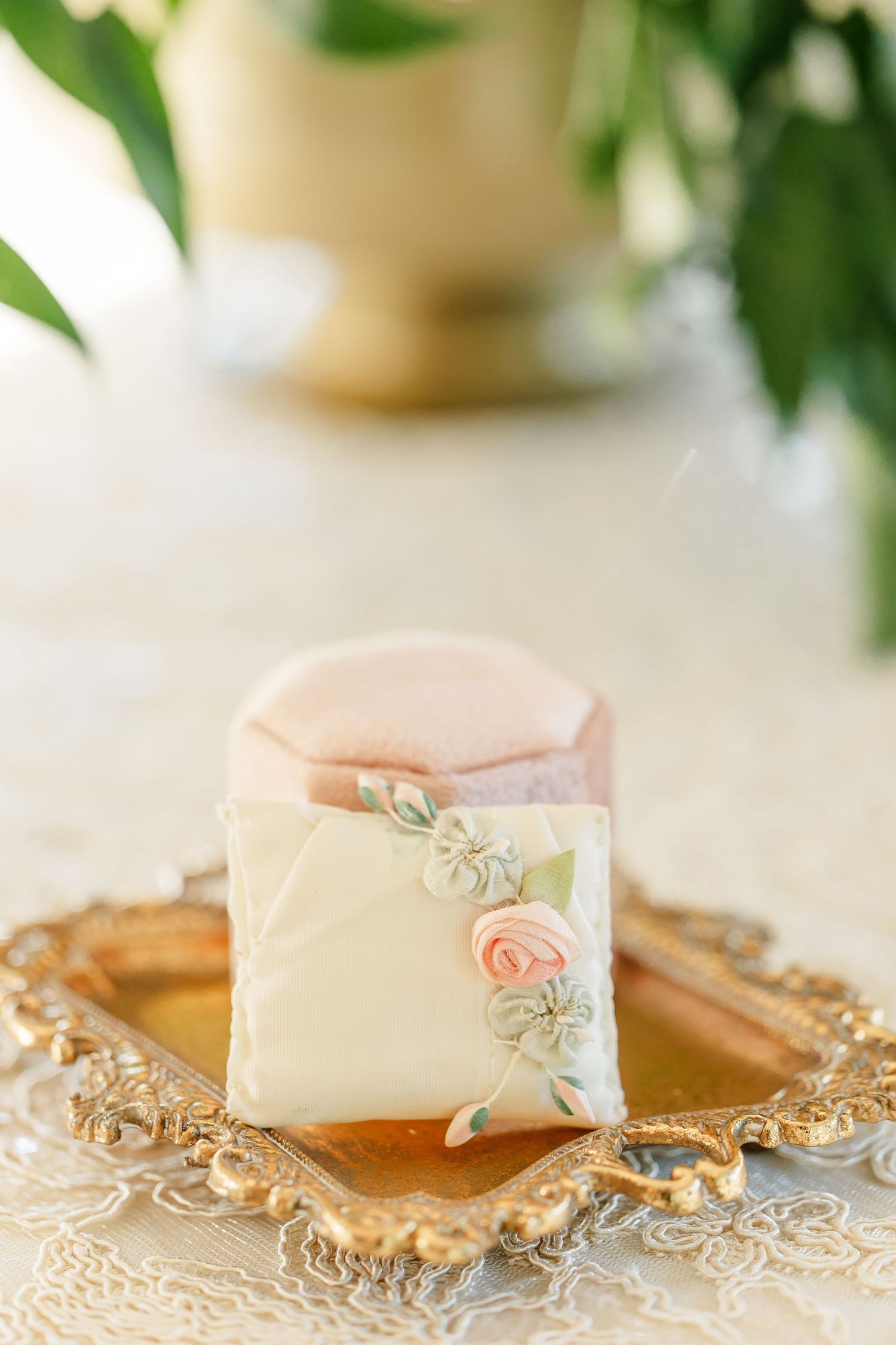 Wedding favor box wrapped in ivory fabric with pink rosebud and green leaf accents on an ornate gold tray atop lace linens.