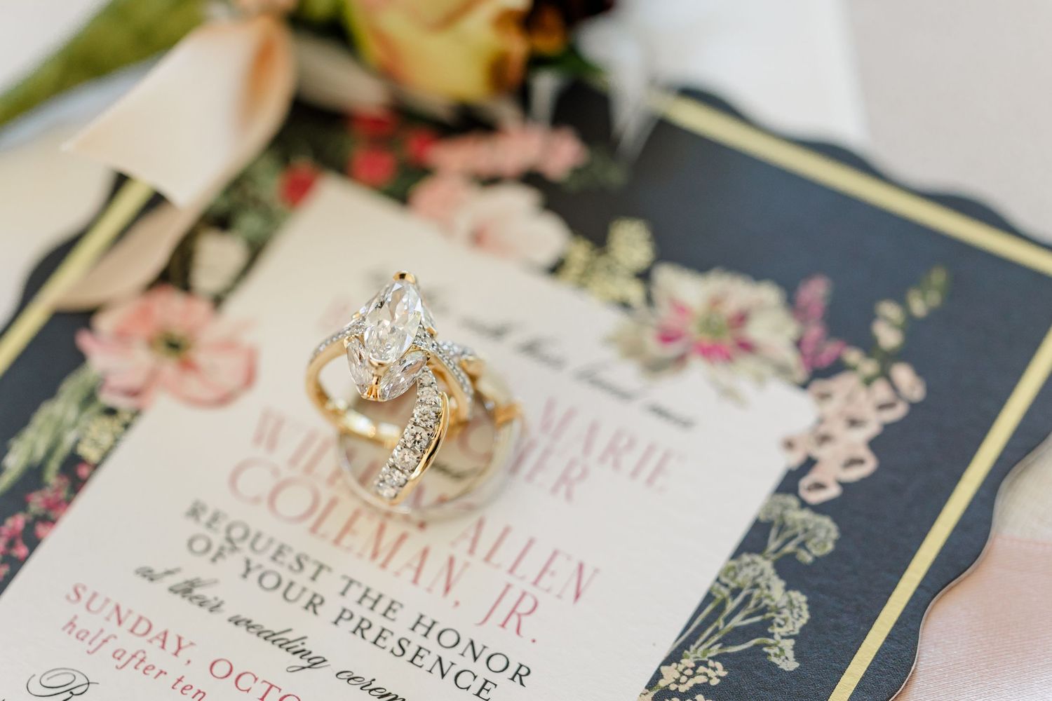 Diamond engagement ring and wedding band resting on a floral wedding invitation with blush roses and a navy and gold border.
