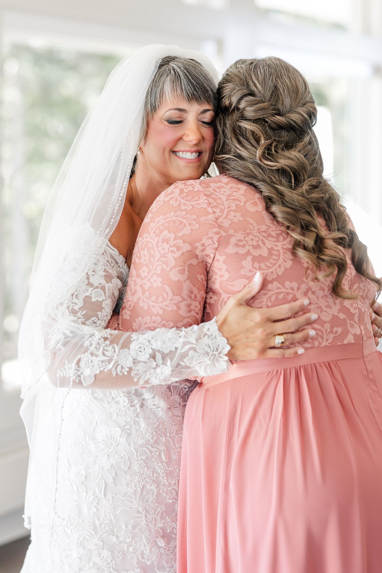 A smiling bride in a lace gown and veil hugs a woman in a blush pink dress with a braided updo before the wedding.