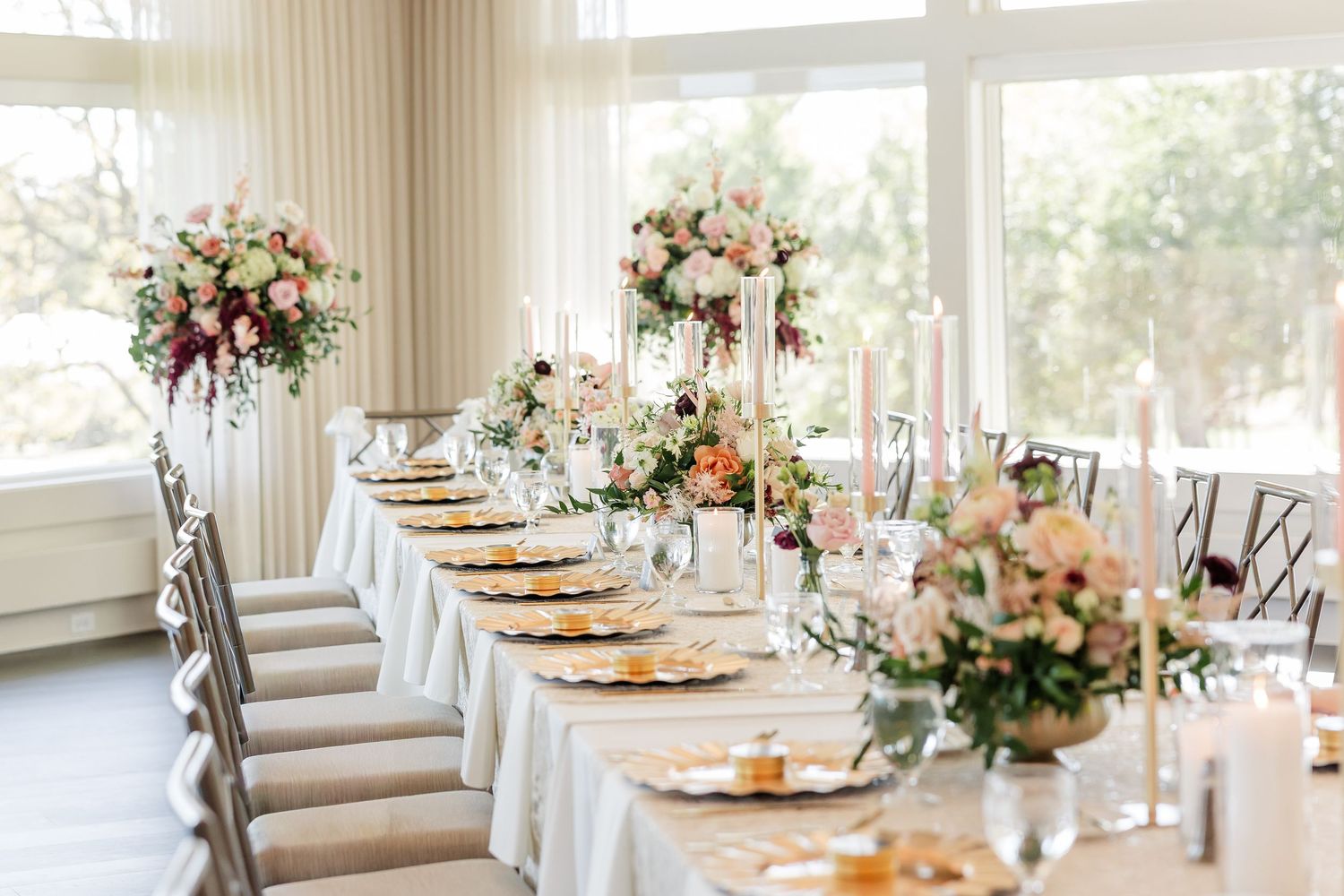 Long elegant wedding reception table with gold chargers, tall candles, and lush pink and white floral centerpieces beside bright windows.