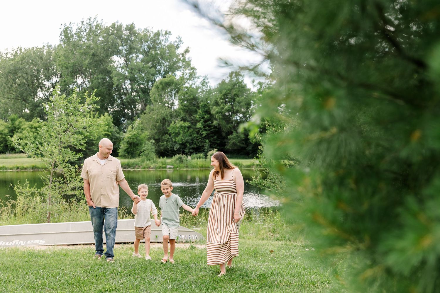 Family of four walking hand-in-hand on green grass near a pond, framed by pine branches on a sunny day.