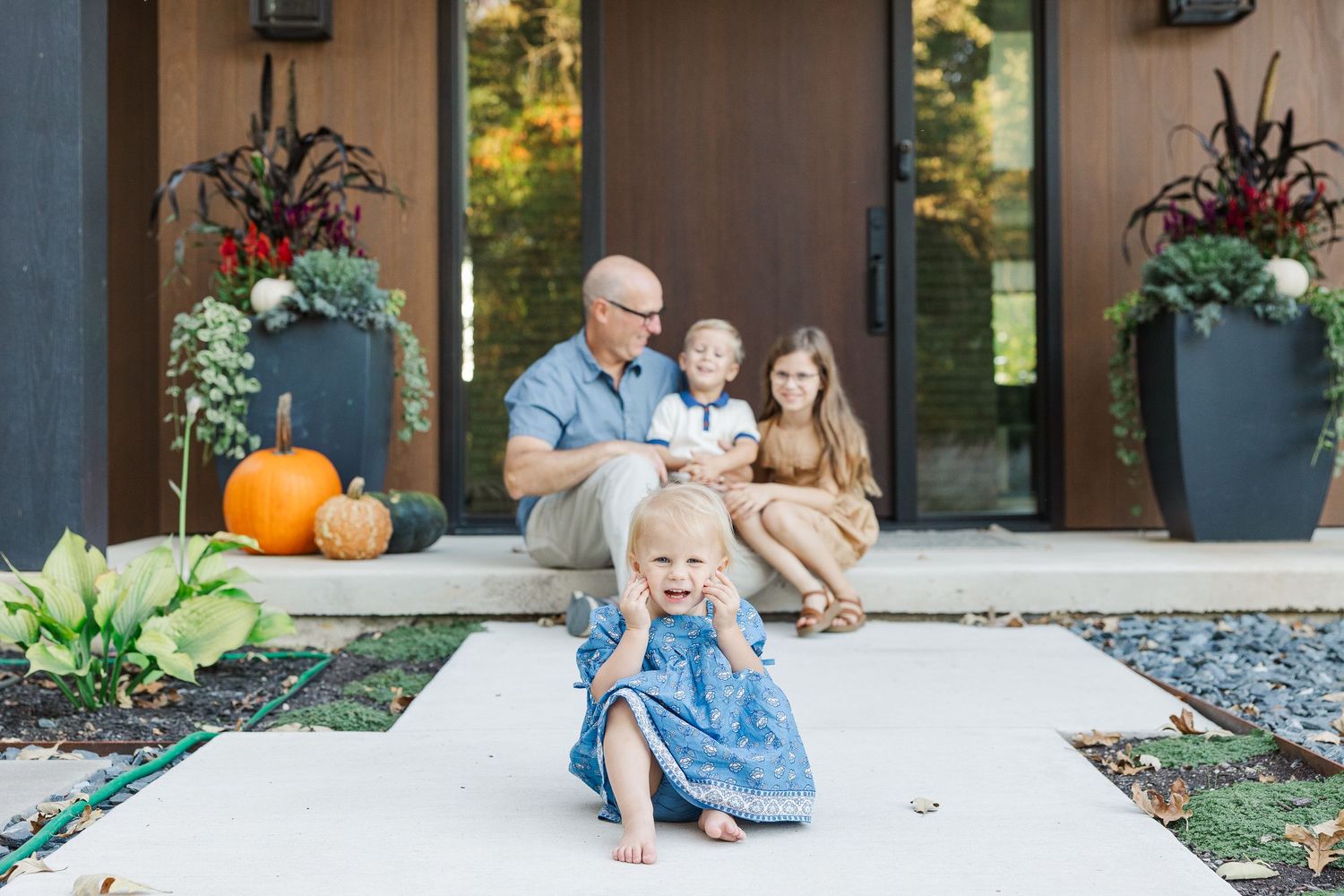 Family sitting together on front porch steps decorated with autumn pumpkins and colorful fall flowers.