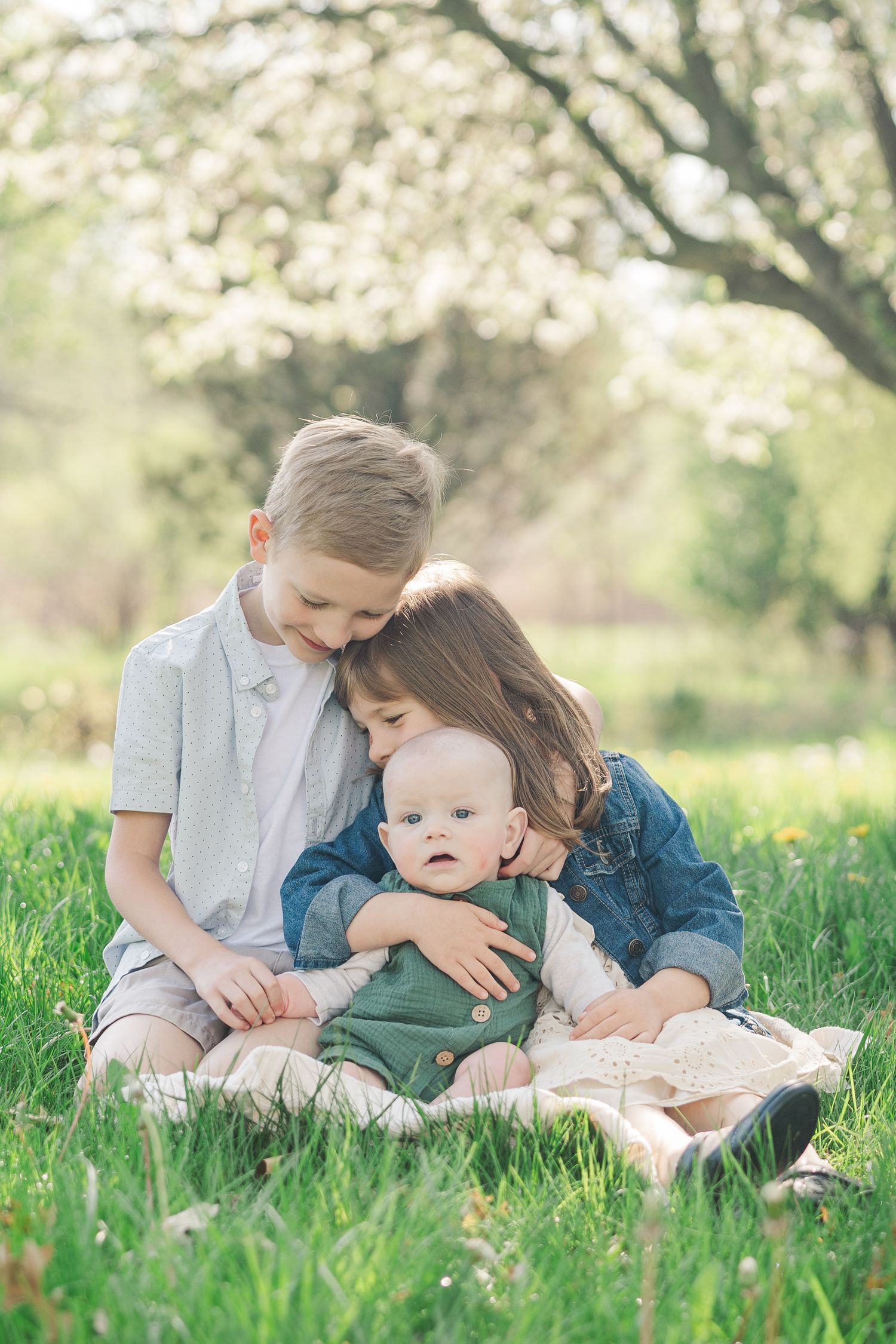 Three siblings share an intimate moment while sitting in grass under flowering trees.