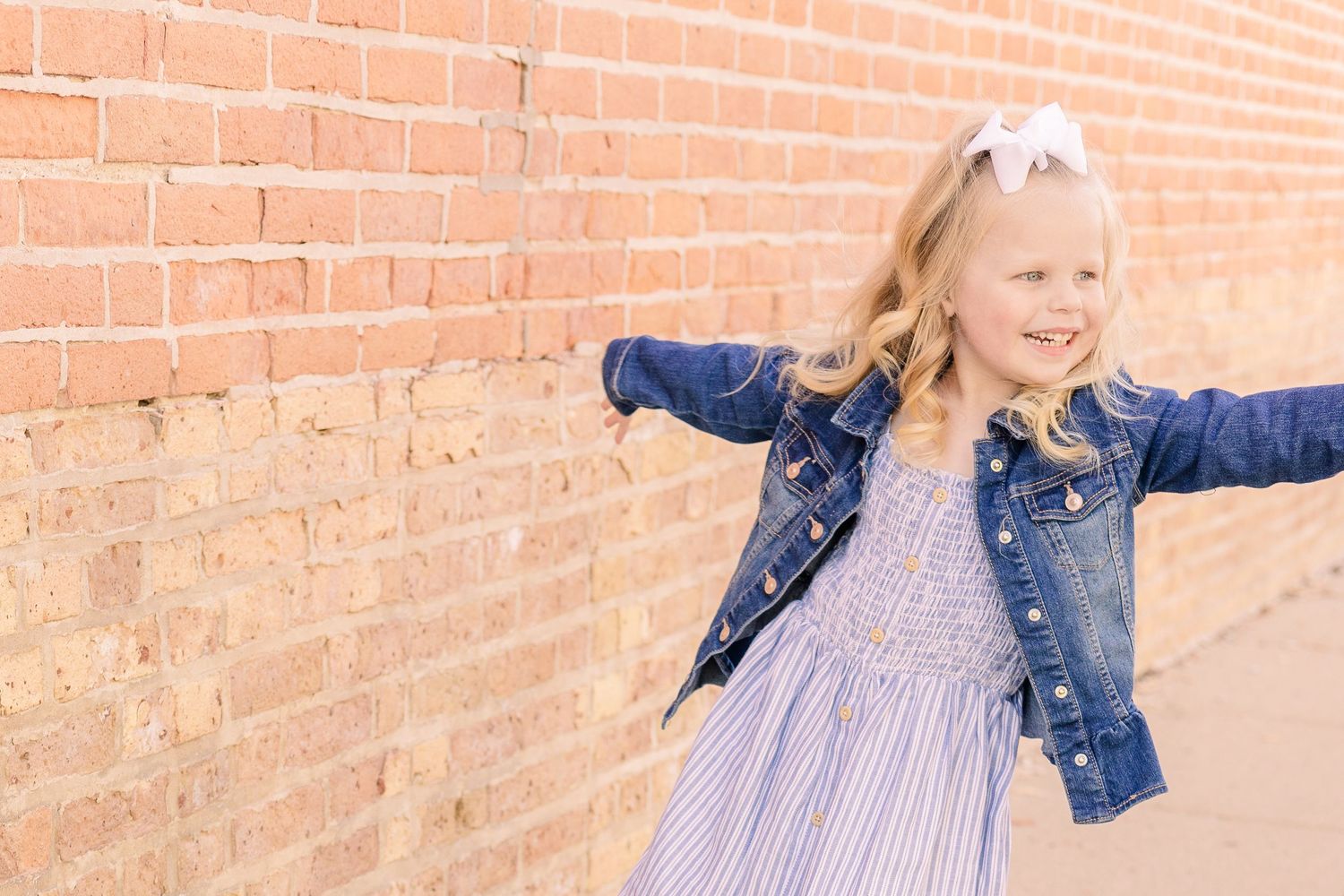 Young child in a denim jacket and striped dress poses playfully against a brick wall.
