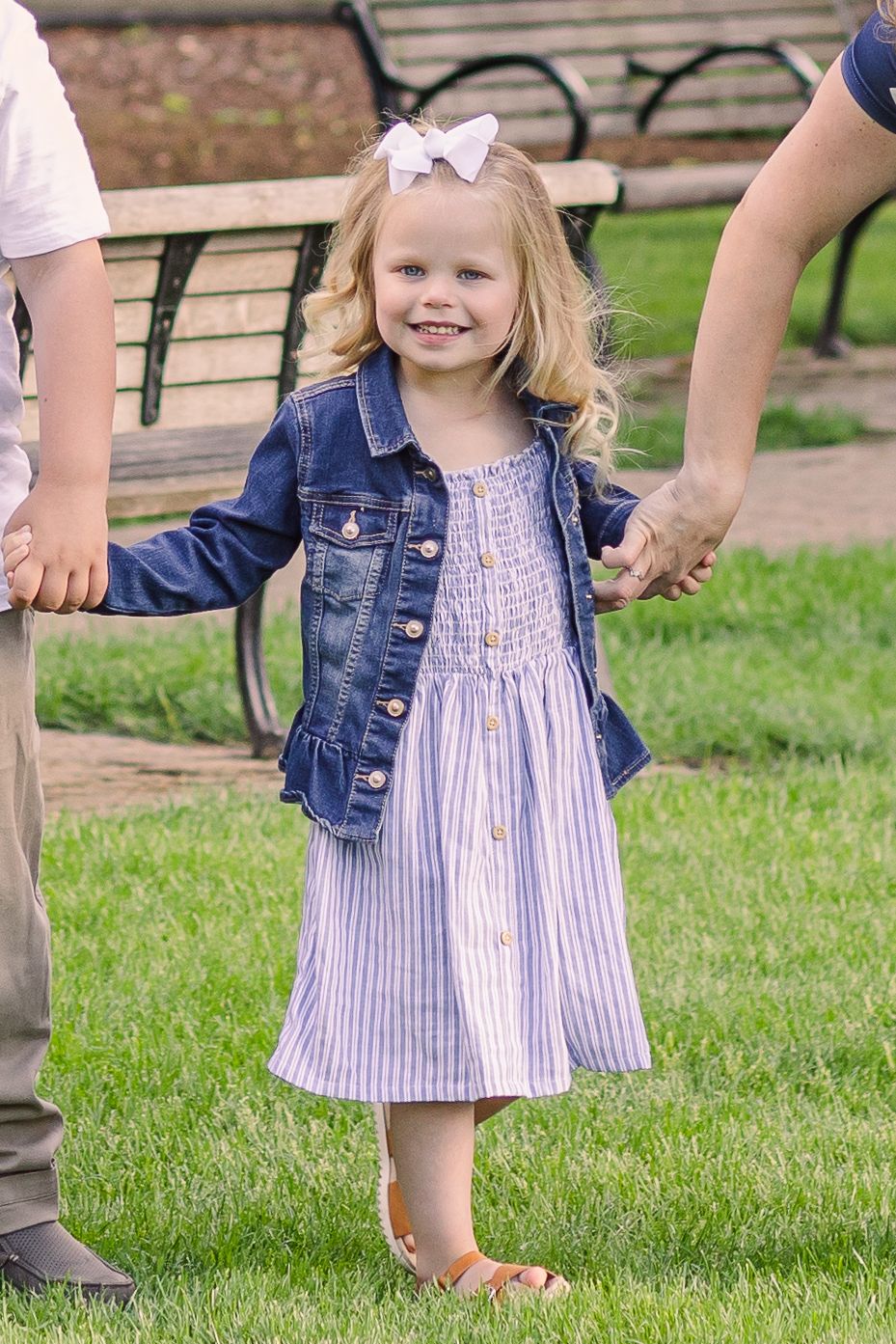Young girl in jean jacket and striped dress smiles while holding hands outdoors.