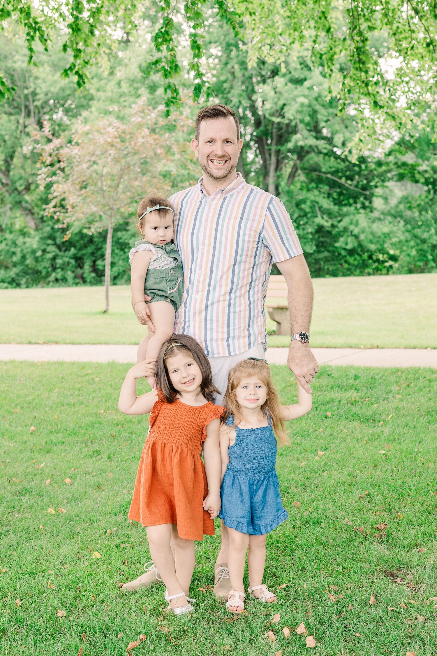 A parent stands with three small children on a grassy lawn surrounded by trees.