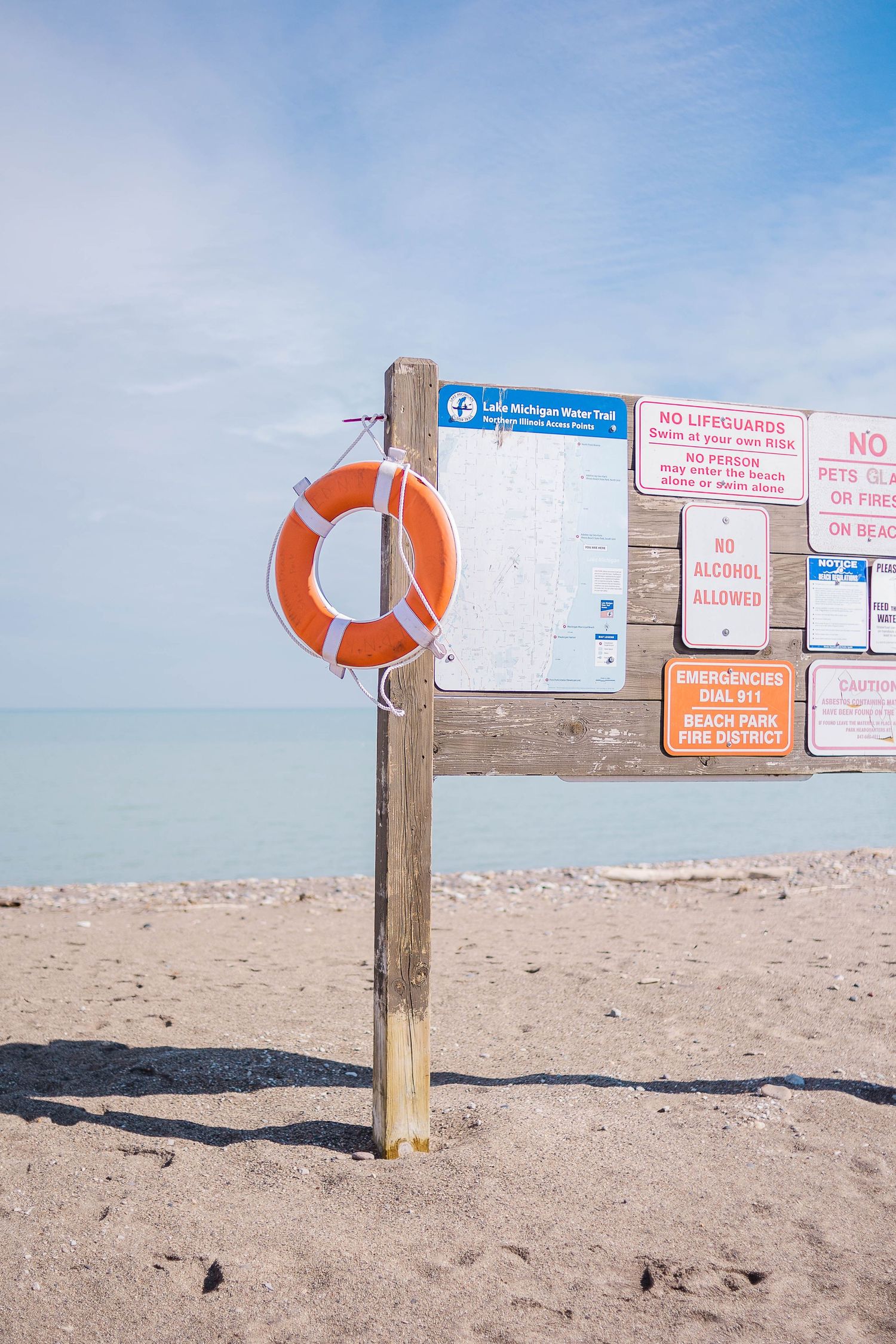 Orange life preserver ring mounted on wooden post with beach safety signs on sandy shoreline.