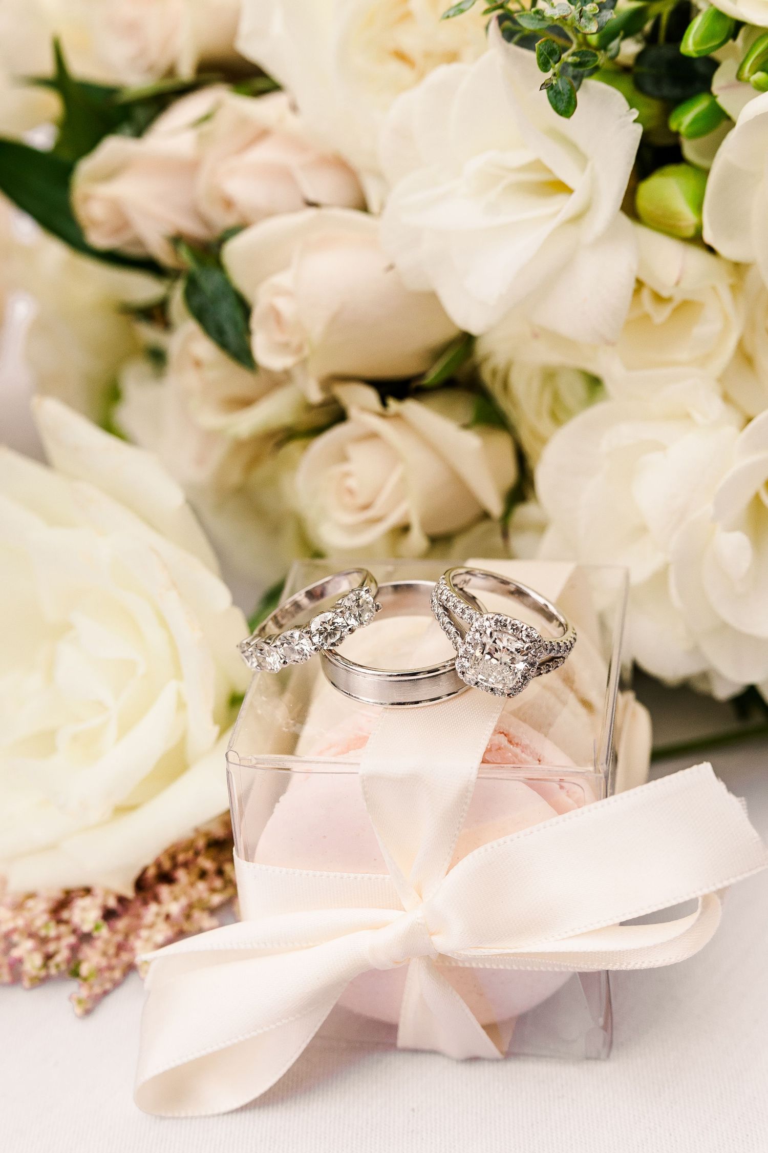 Wedding rings resting on a clear box with pink bow surrounded by white roses.