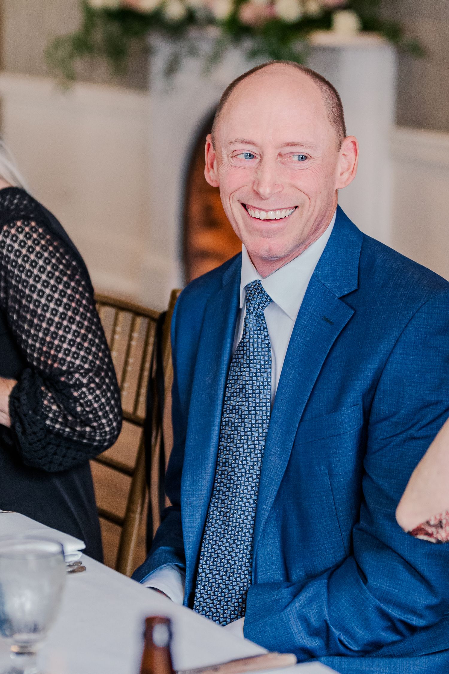 Wedding guest in a navy blue suit and patterned tie smiling at reception dinner.
