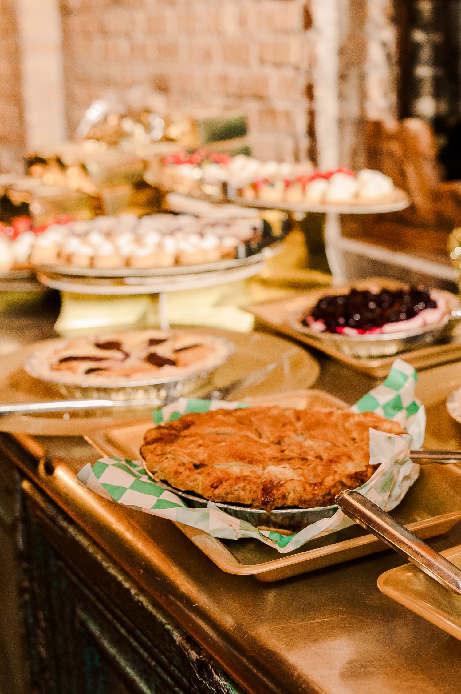 Dessert table display with pies and pastries on tiered stands against a rustic brick wall.