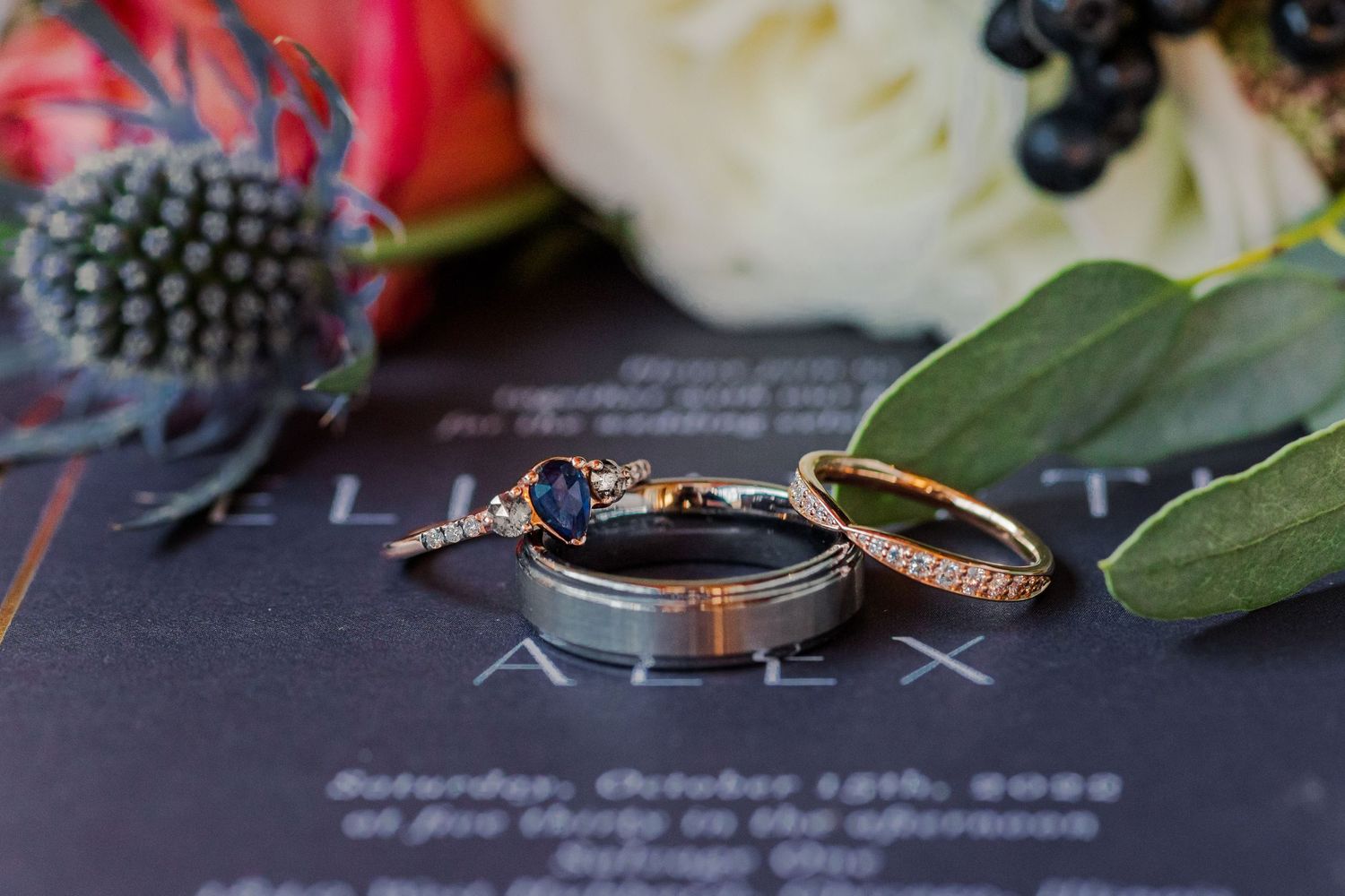 Wedding ring set displayed on dark navy invitation with floral accents and greenery.