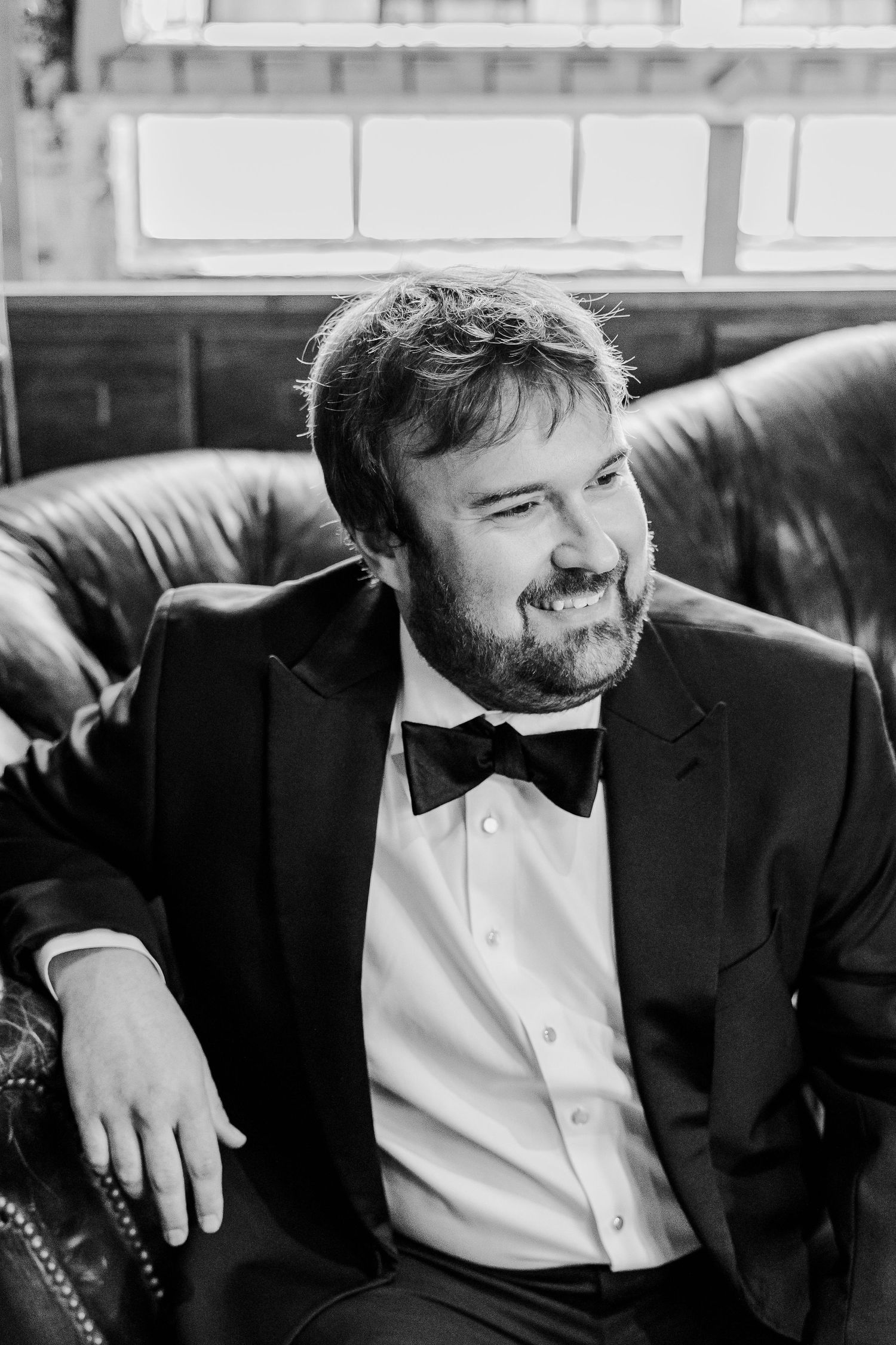 A groomsman wearing a black tuxedo and bowtie sits on a leather couch.