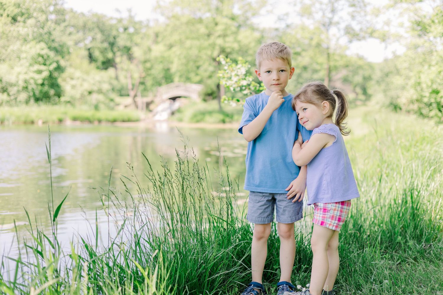 Two young children standing by a pond looking at something in the water.