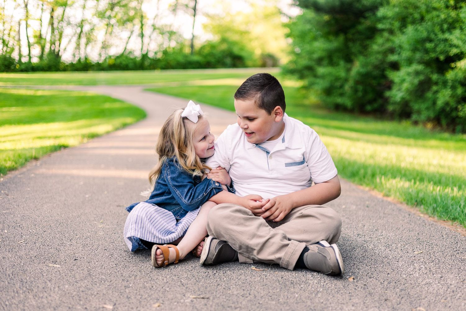 Two people sit together on a winding park path surrounded by green grass and trees.