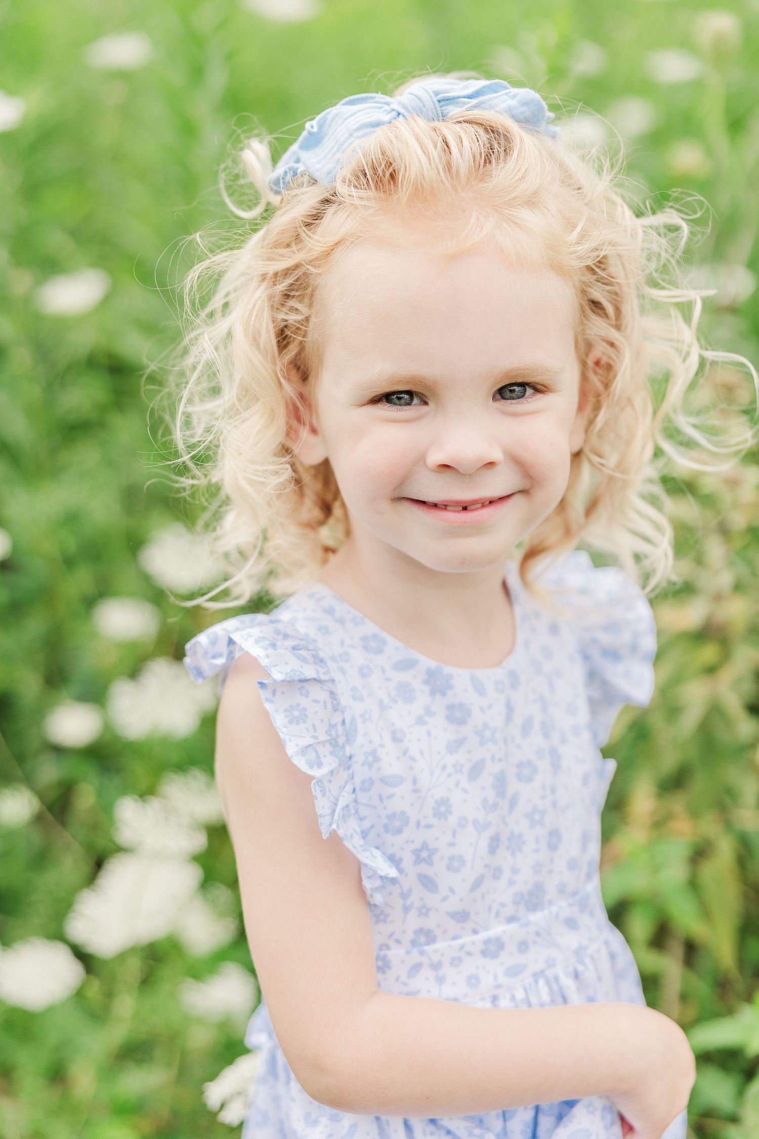 A close-up portrait of a young child in a blue floral dress against a green summer backdrop.