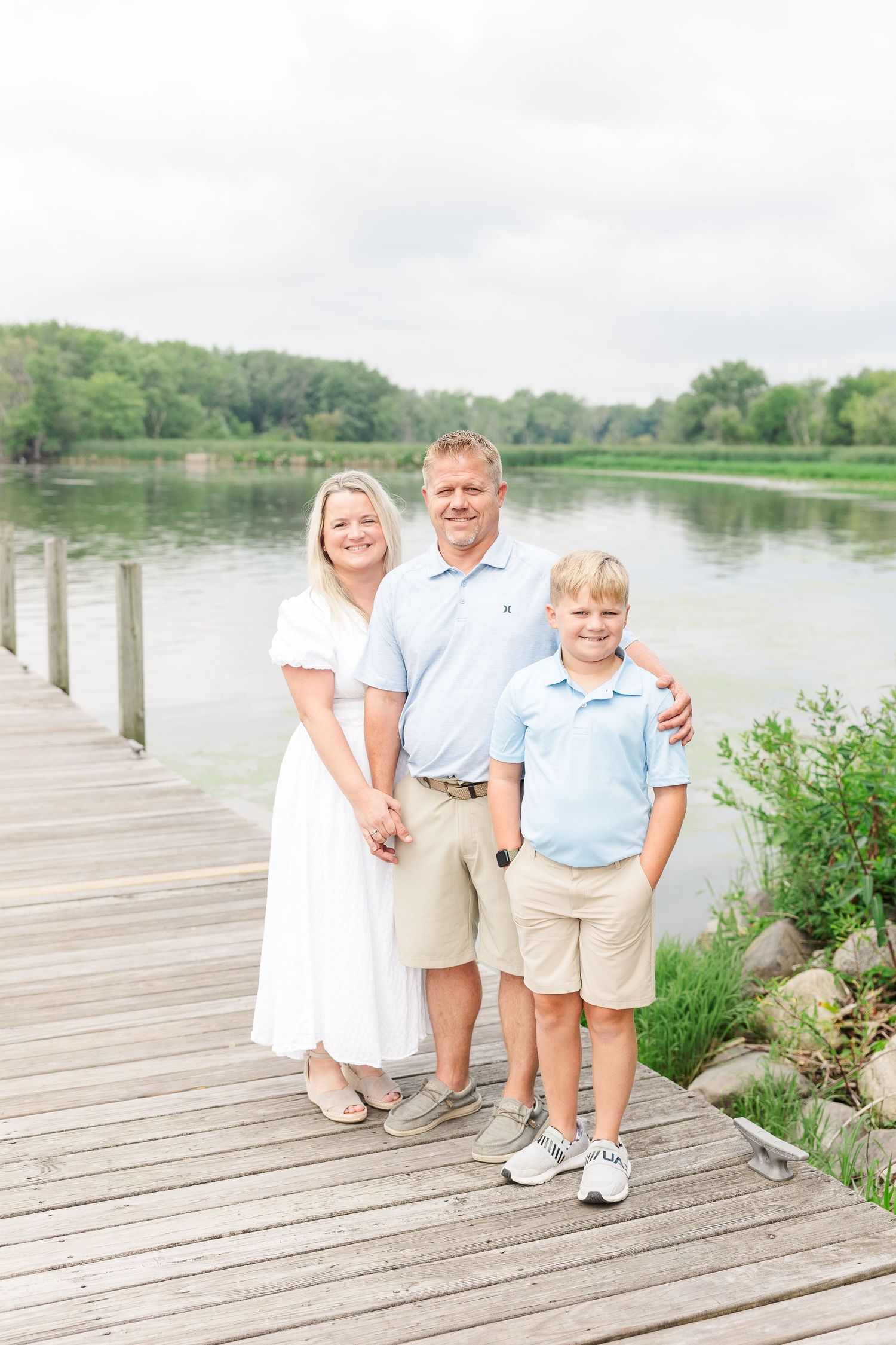 Three people pose on a wooden pier beside a tranquil lake with lush greenery in the background.