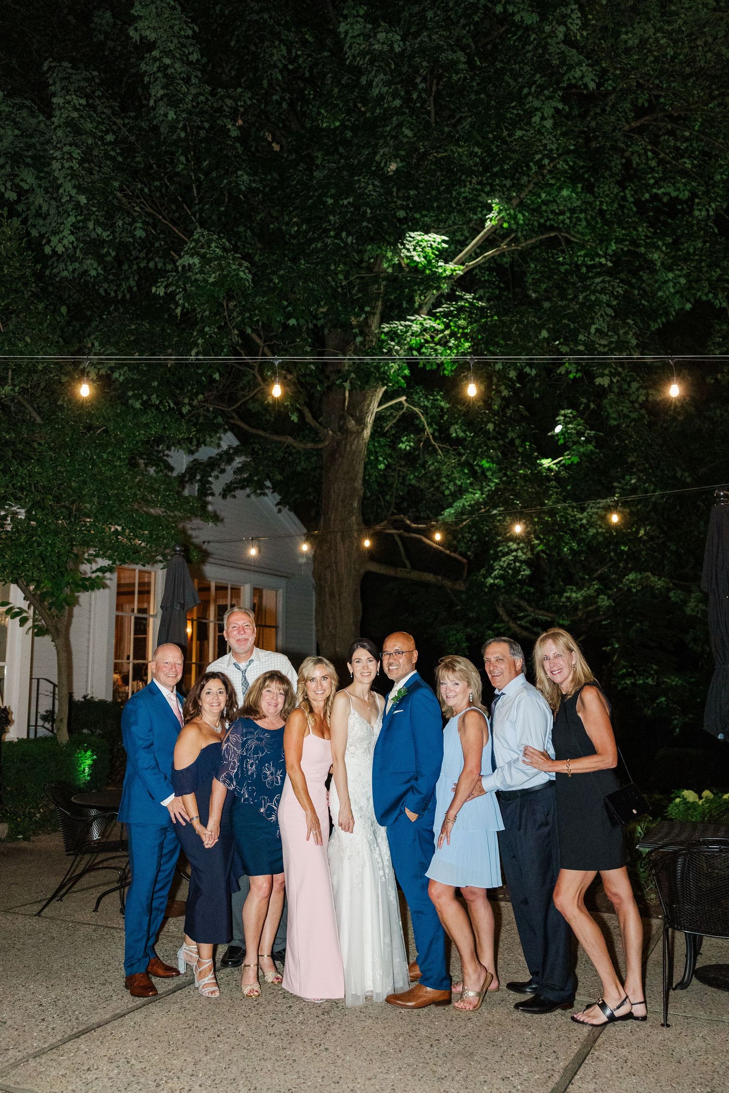 A wedding party poses under string lights and trees during an evening outdoor photo session.