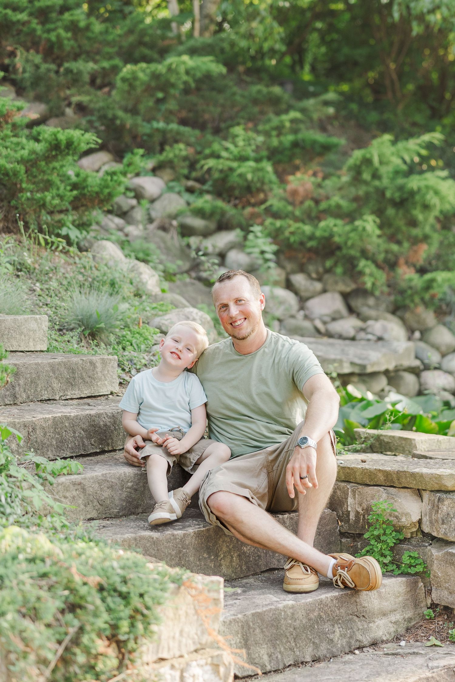 Two people sitting together on stone garden steps surrounded by greenery and landscaping on a sunny day.