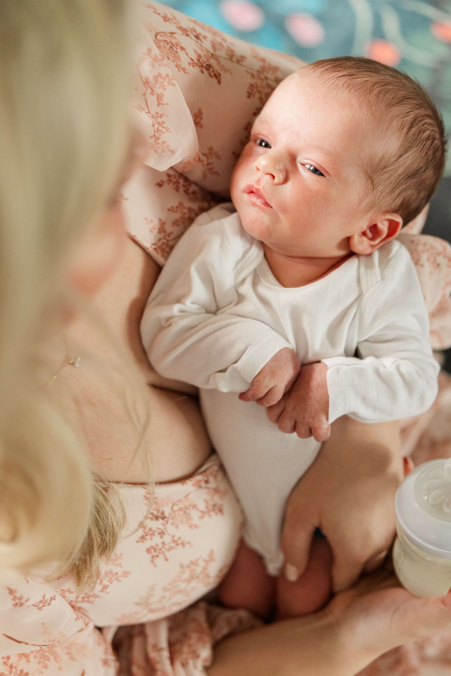 Newborn baby in white onesie gazing at blonde mother while being held with a bottle nearby.