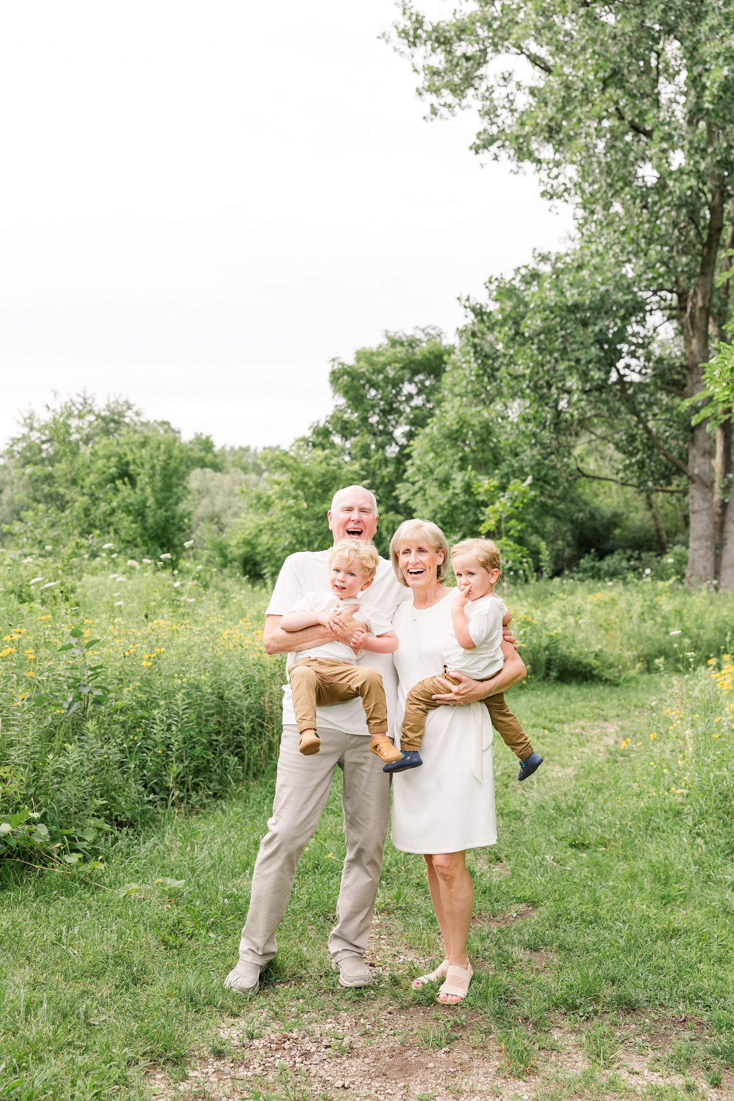 Happy grandparents holding two toddler grandchildren outdoors in a green park setting on a cloudy day.