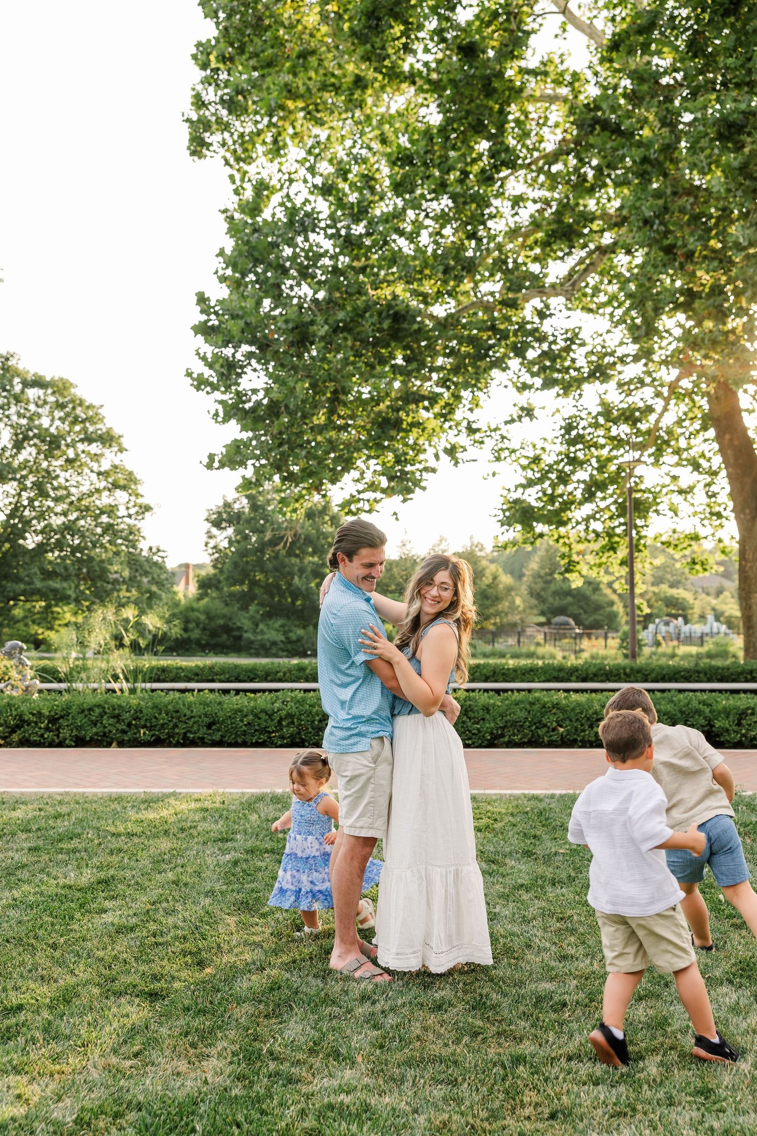 Parents embrace on green grass while two young children play around them in a sunlit park.