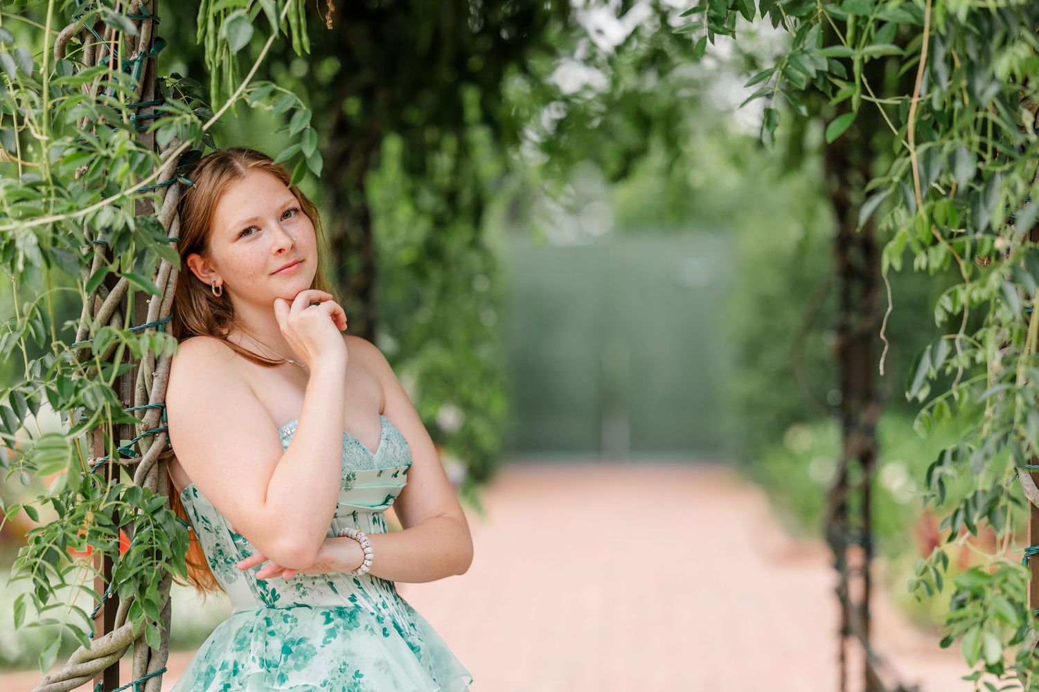 Young woman in floral dress posing thoughtfully in a lush green garden archway on a sunny day.