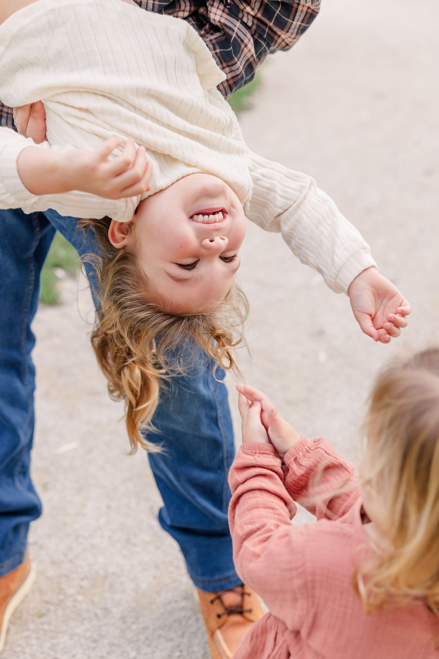 A laughing child is playfully held upside down by an adult while a sibling in pink reaches out.