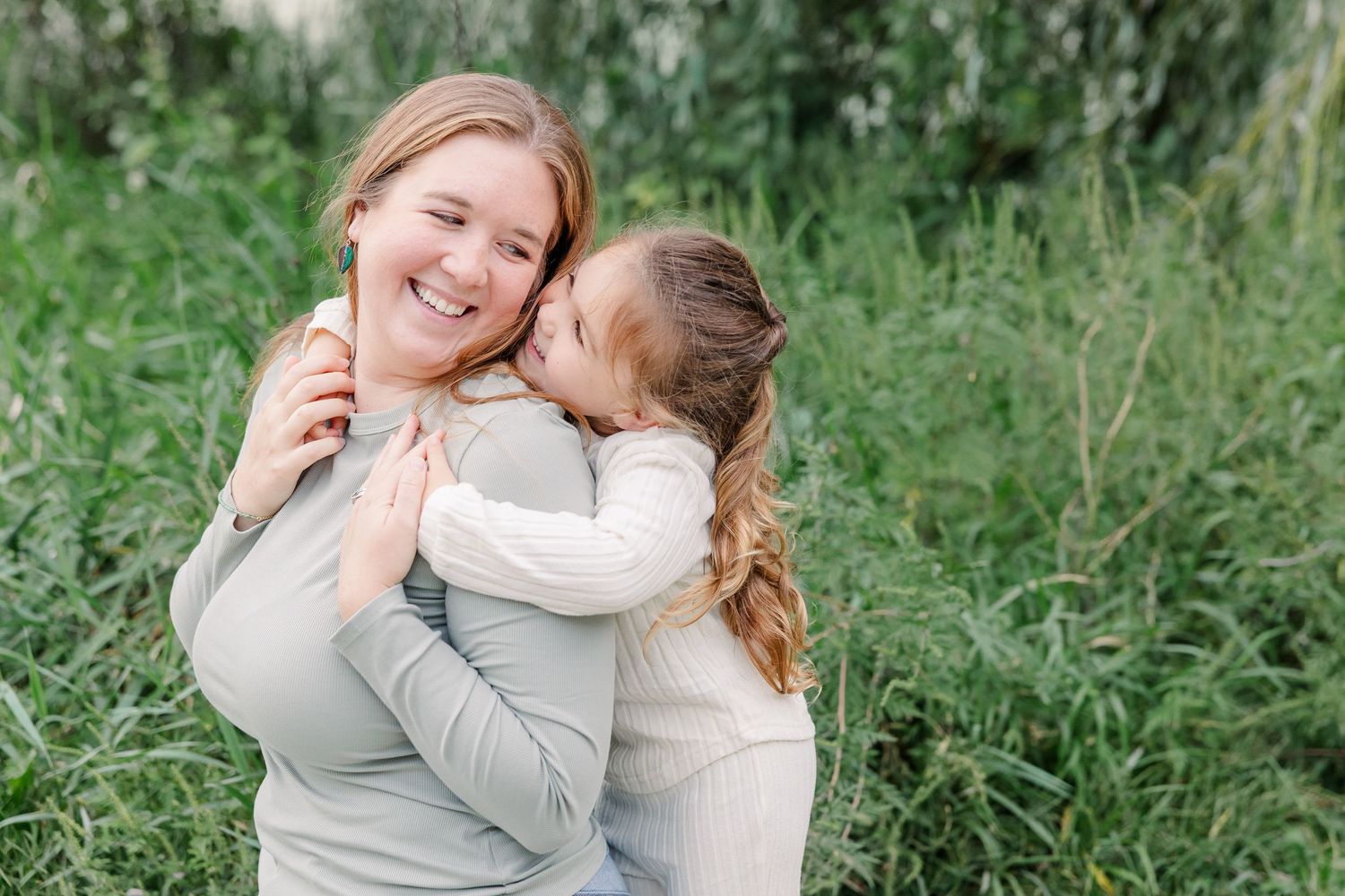 A young girl kisses her smiling mother on the cheek while embracing outdoors in a lush green field.