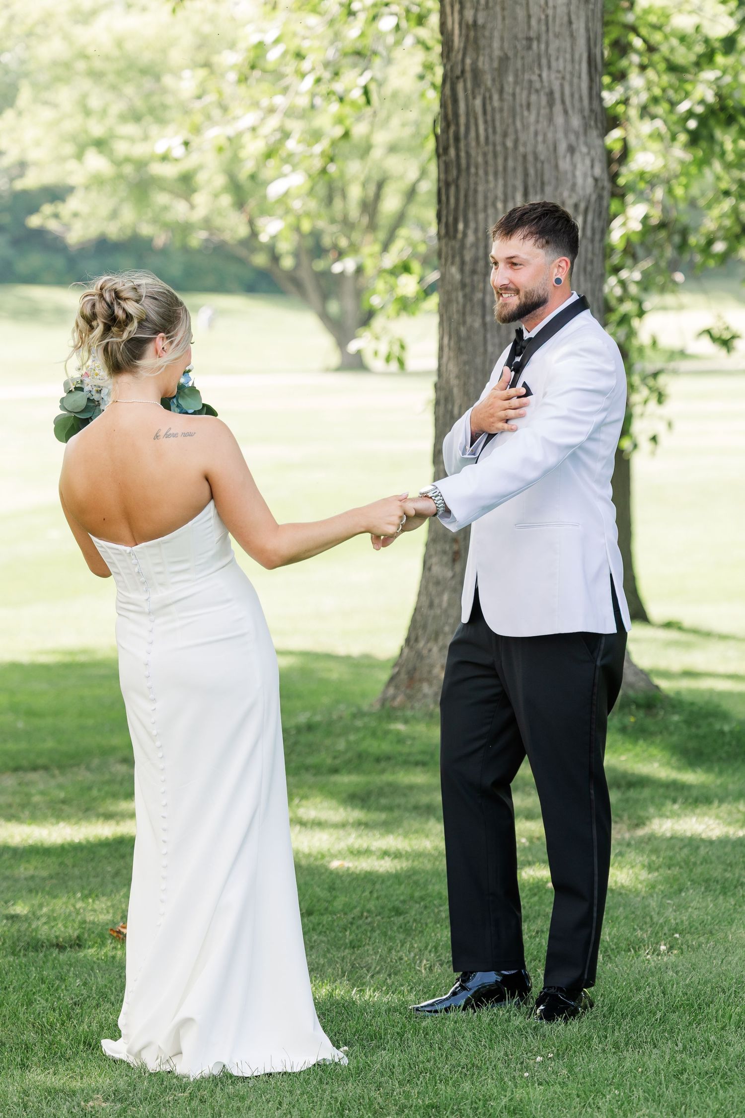 Smiling groom in white tuxedo jacket holds bride's hands during outdoor first look on wedding day.