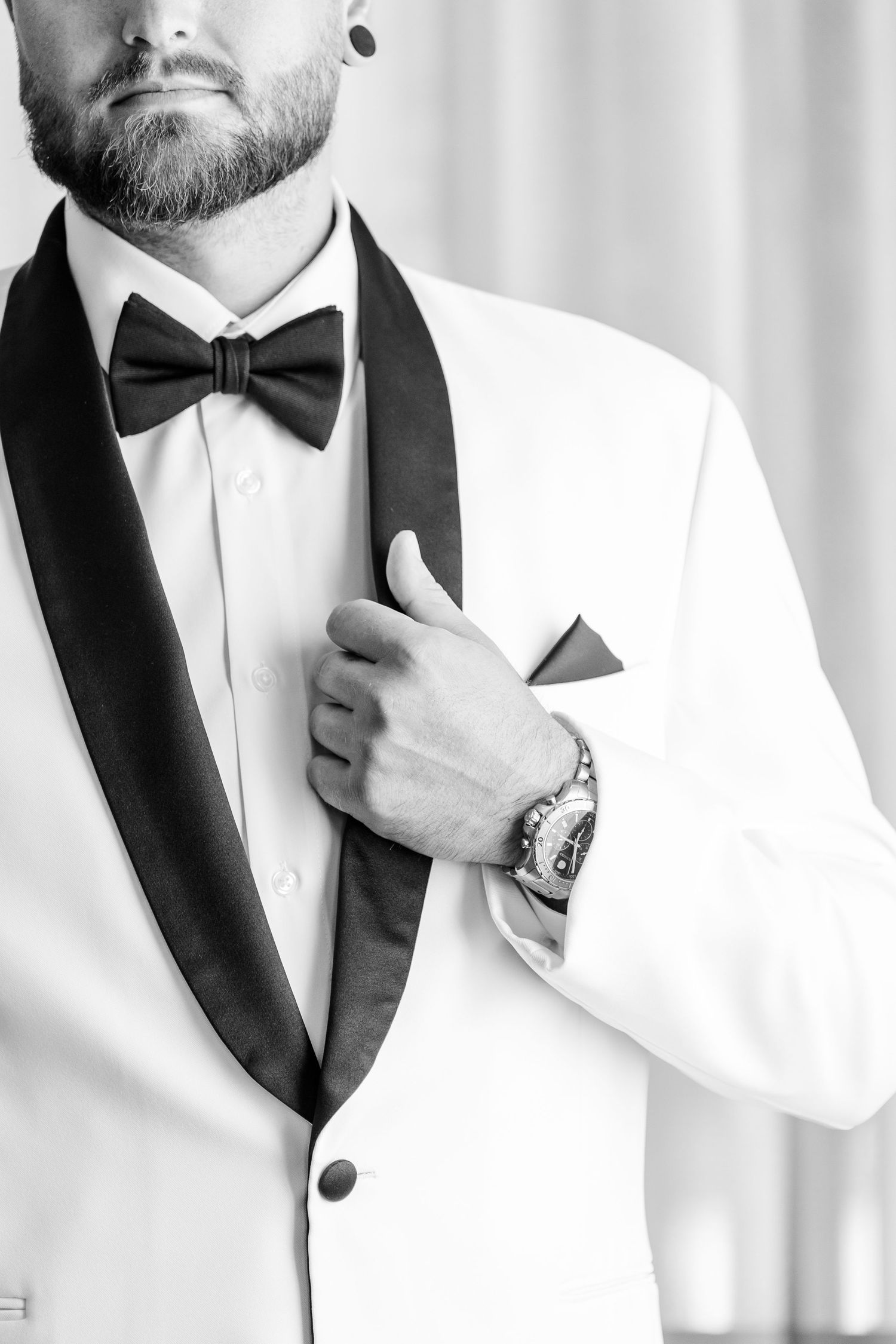 Groom in white tuxedo jacket with black bow tie and pocket square adjusting lapel before wedding ceremony.